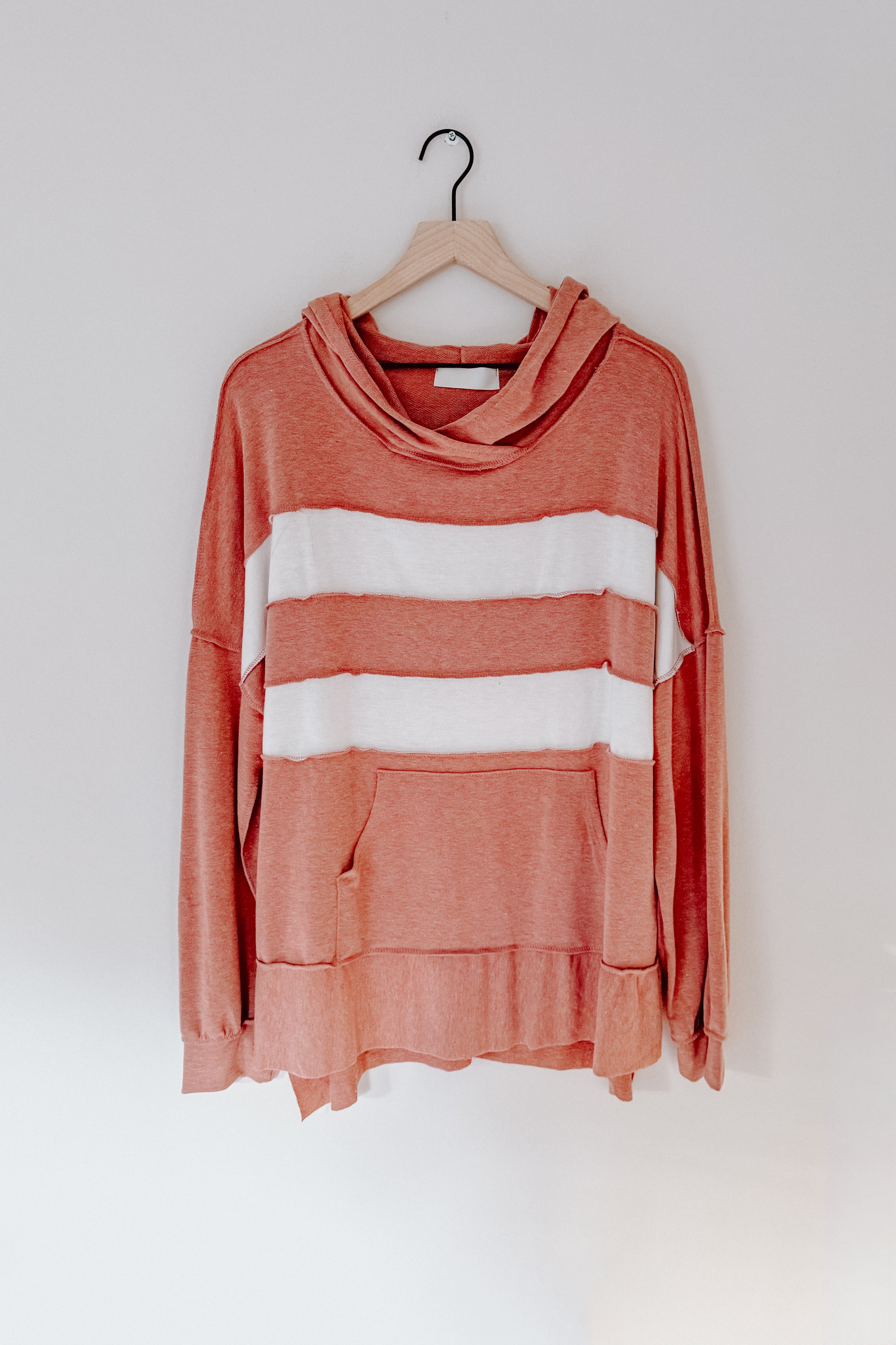 Rust-colored hooded pullover with loose fit featuring two wide white horizontal stripes and a front pocket hanging on wooden hanger