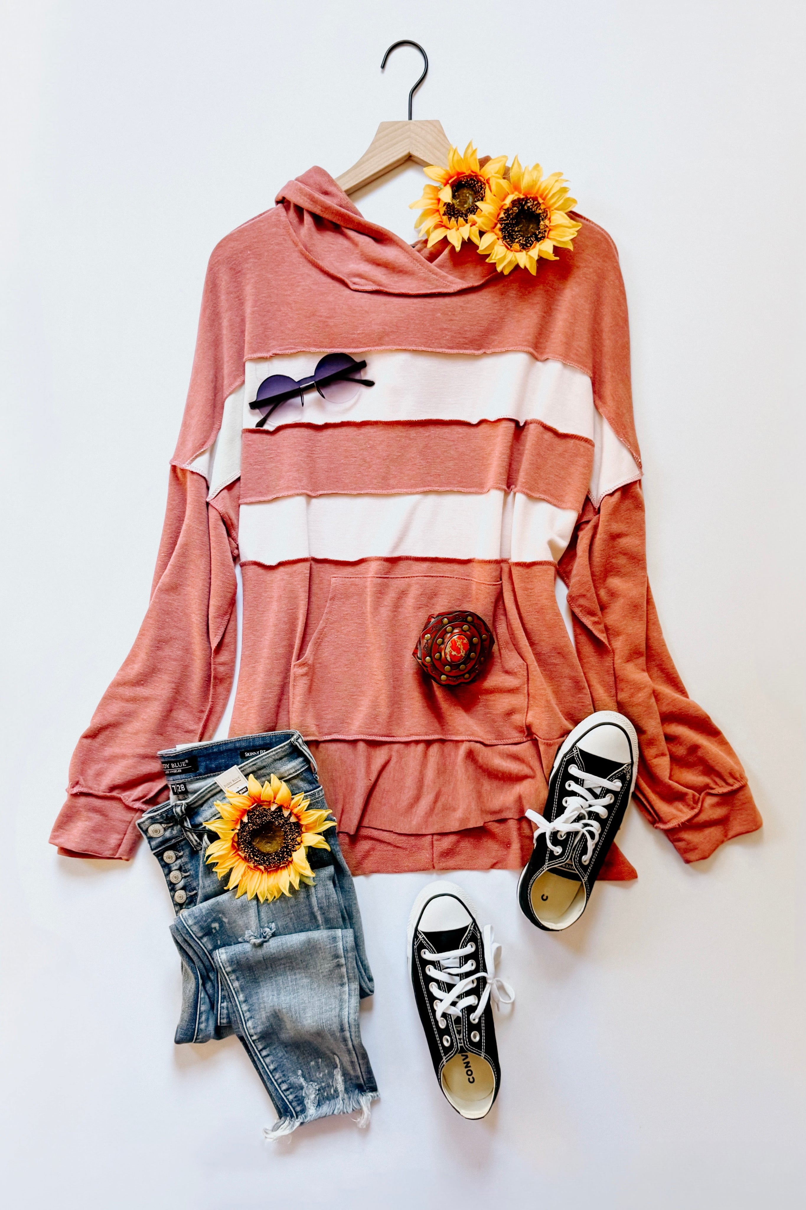 Rust-colored hoodie with white stripes paired with distressed blue jeans and black canvas sneakers flat lay with sunflowers and sunglasses