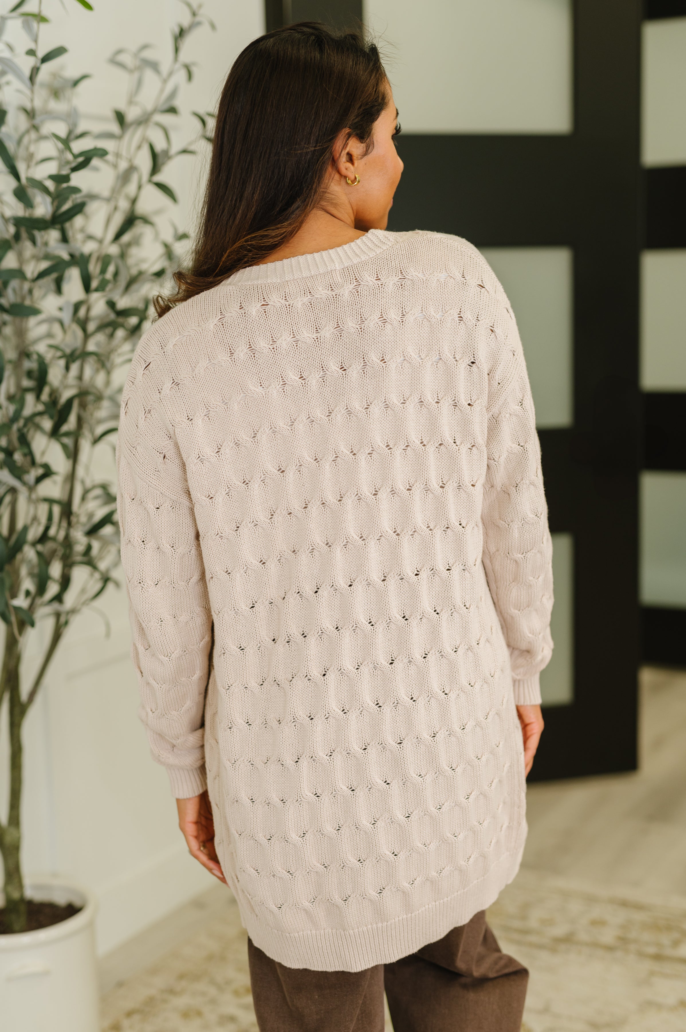 Woman wearing a light beige cable knit cardigan with long sleeves seen from the back indoors