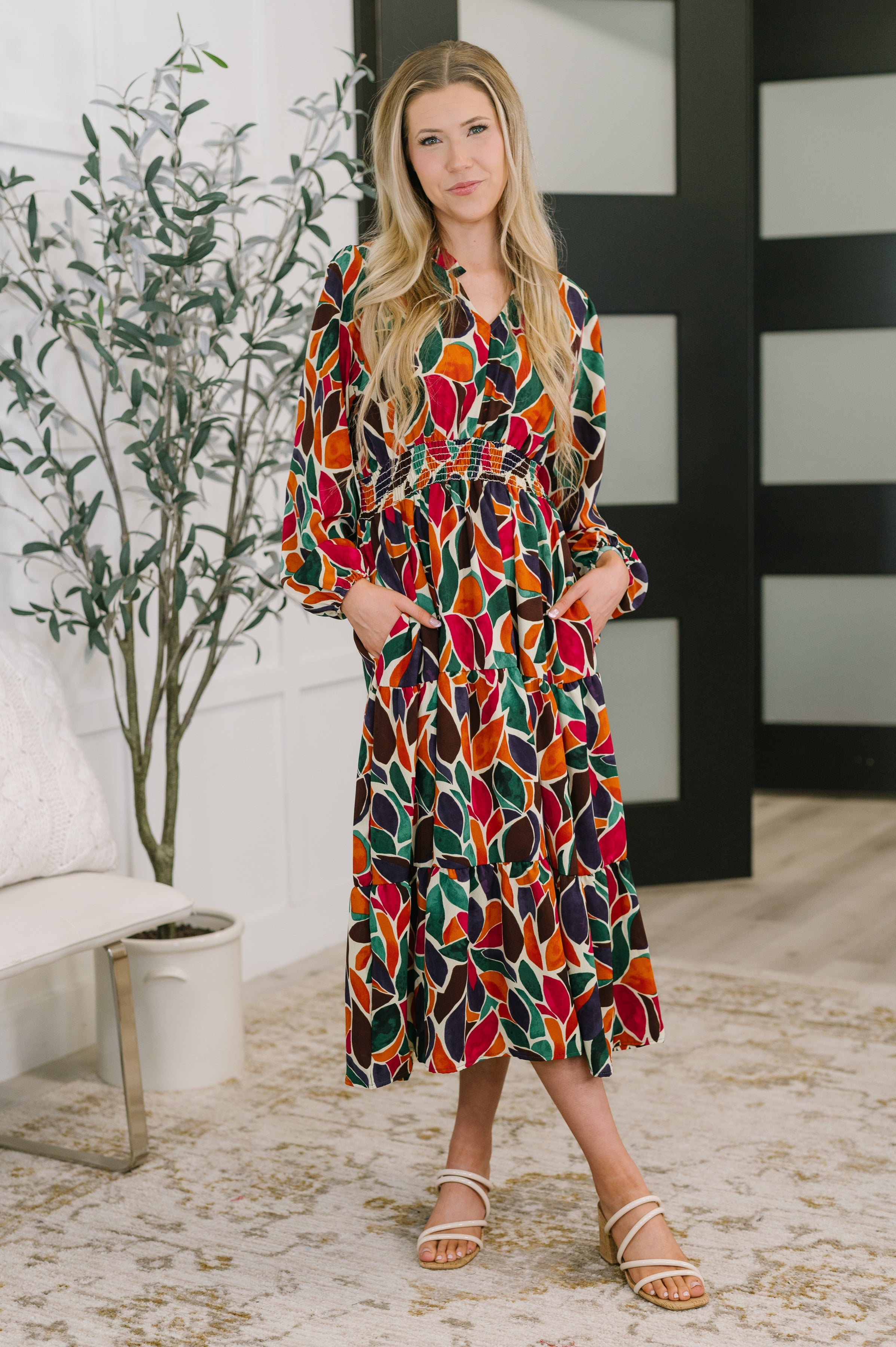 Woman wearing a colorful long sleeve tiered dress with an abstract leaf pattern and pockets indoors
