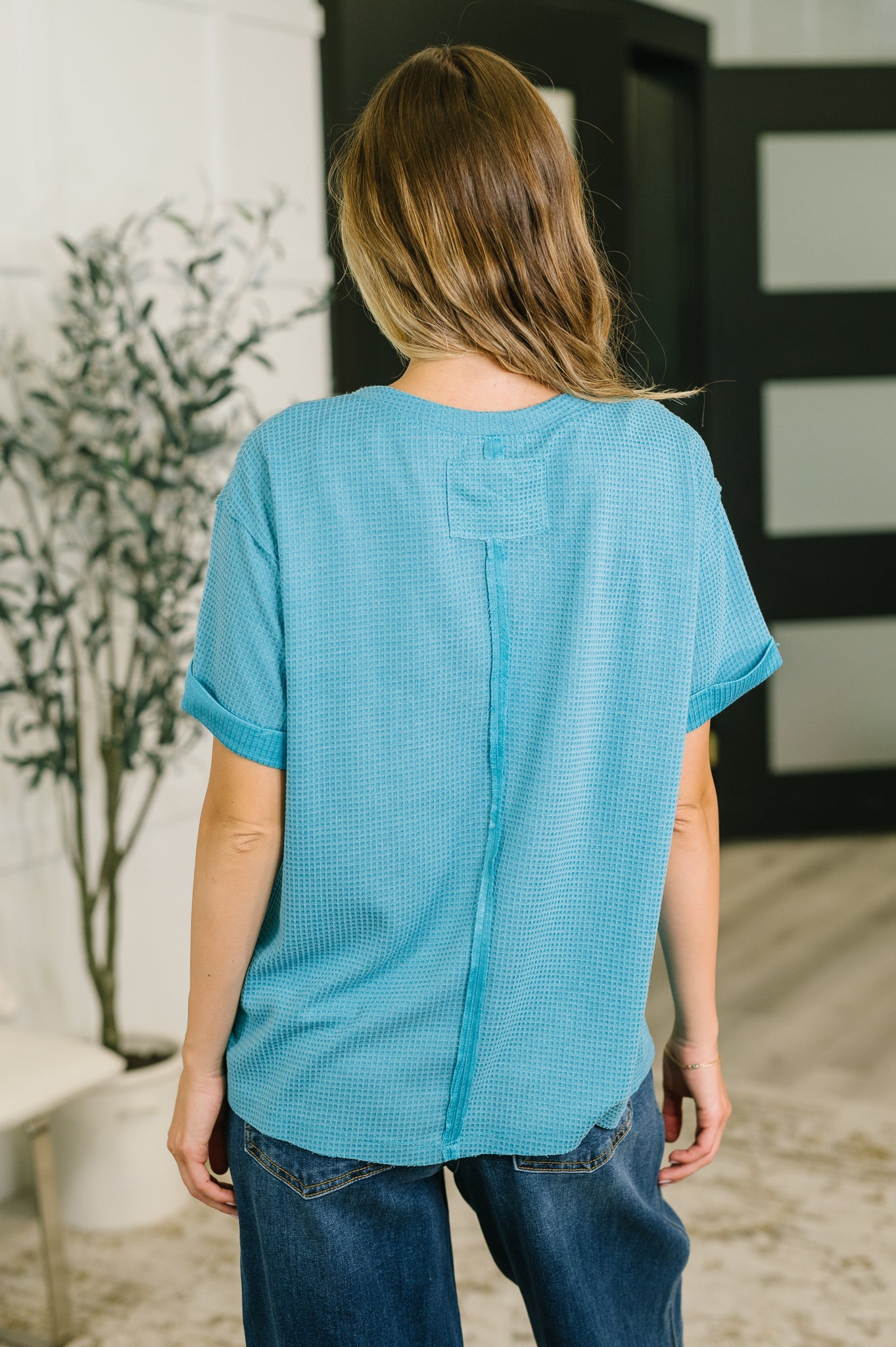 Back view of woman wearing a teal waffle knit relaxed tee with rolled sleeves paired with loose blue jeans indoors