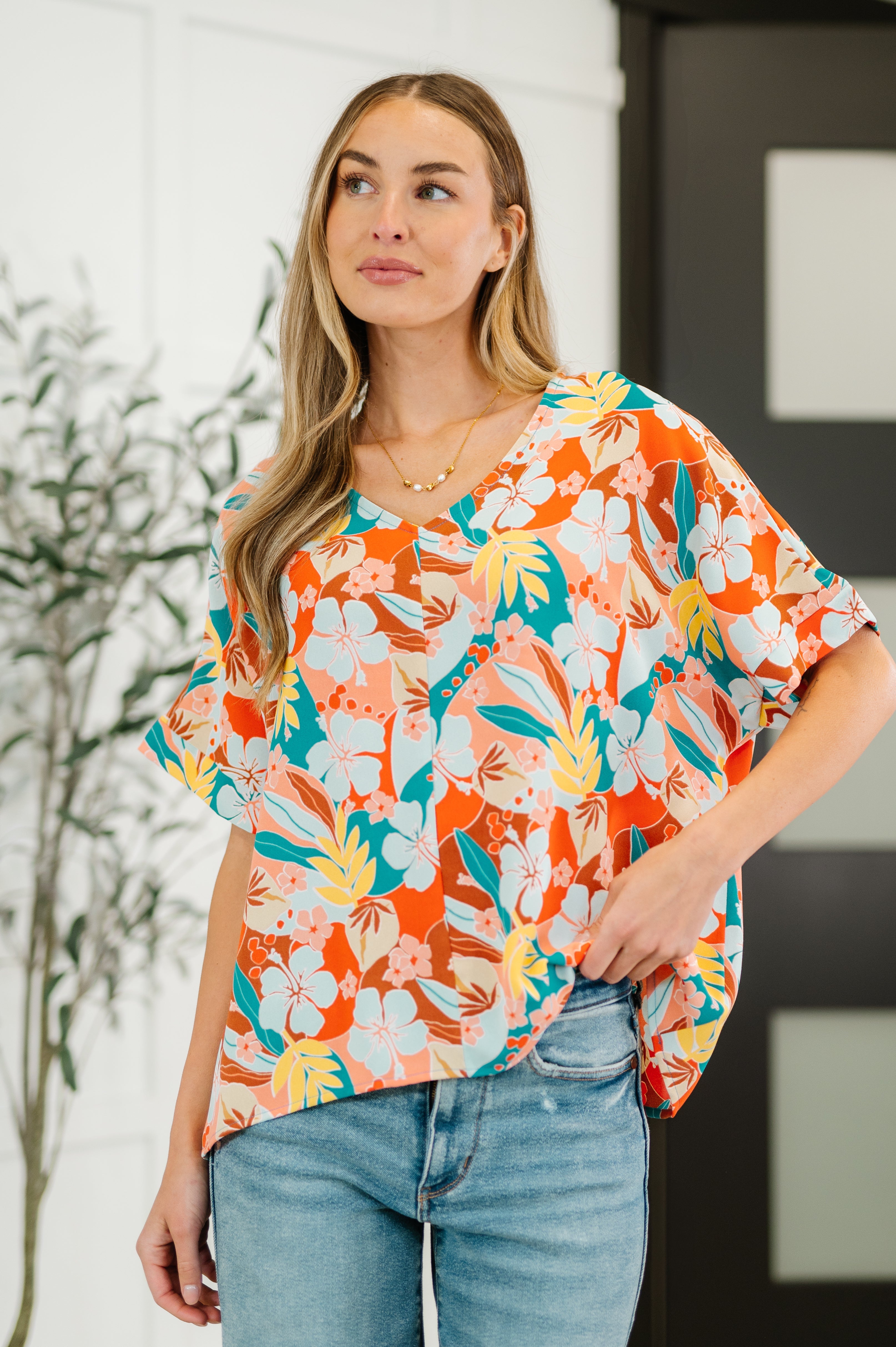 Woman wearing loose orange floral V-neck top paired with light-wash jeans and layered gold necklace indoors