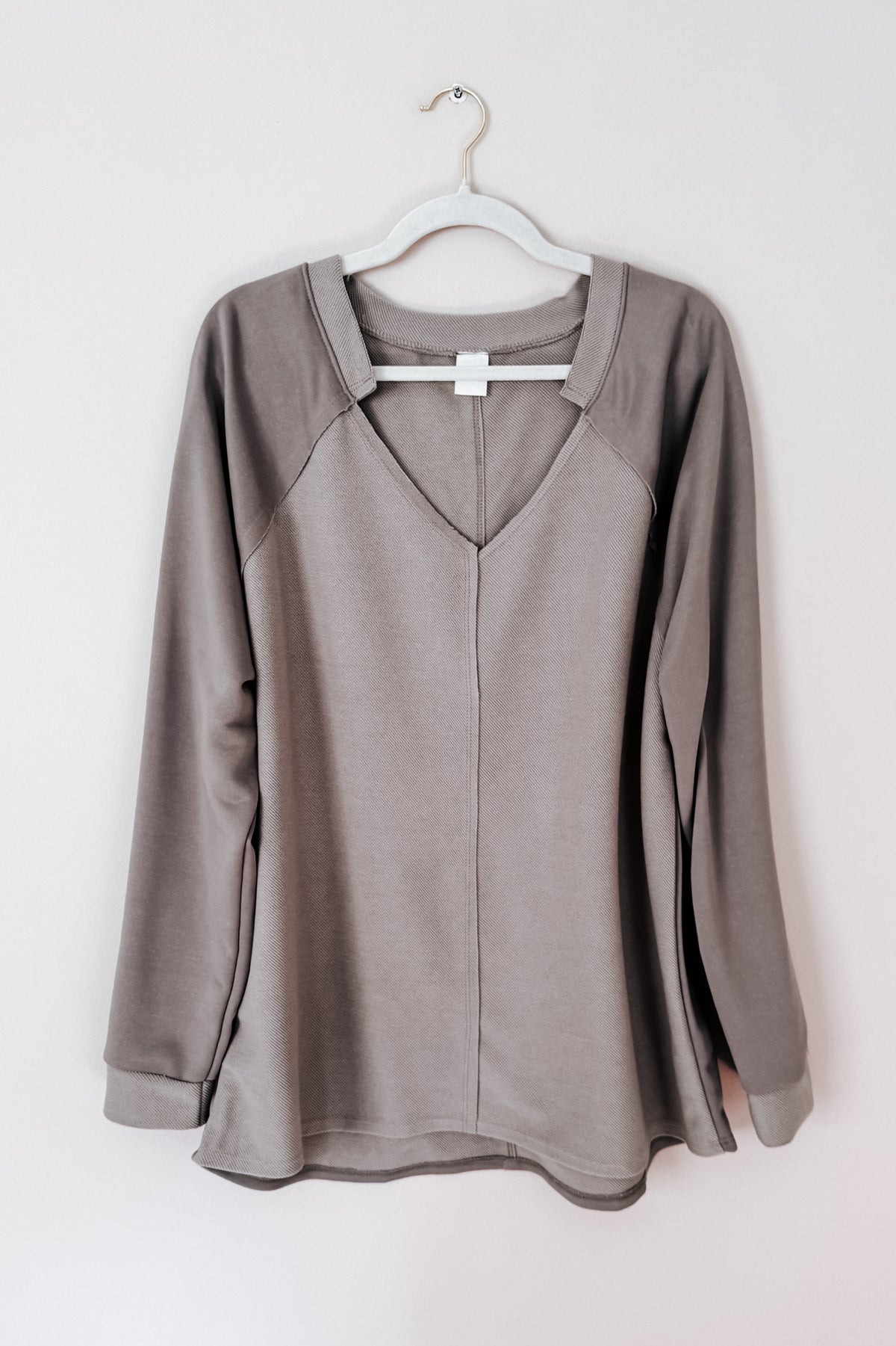 Beige textured knit top with a notched V neckline raglan sleeves and raw edges hanging from a white hanger