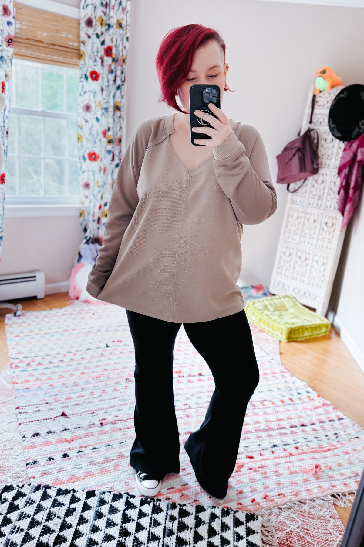 Woman taking a mirror selfie in a beige textured knit top with a notched V neckline and raglan sleeves black flared pants and sneakers in a colorful bedroom