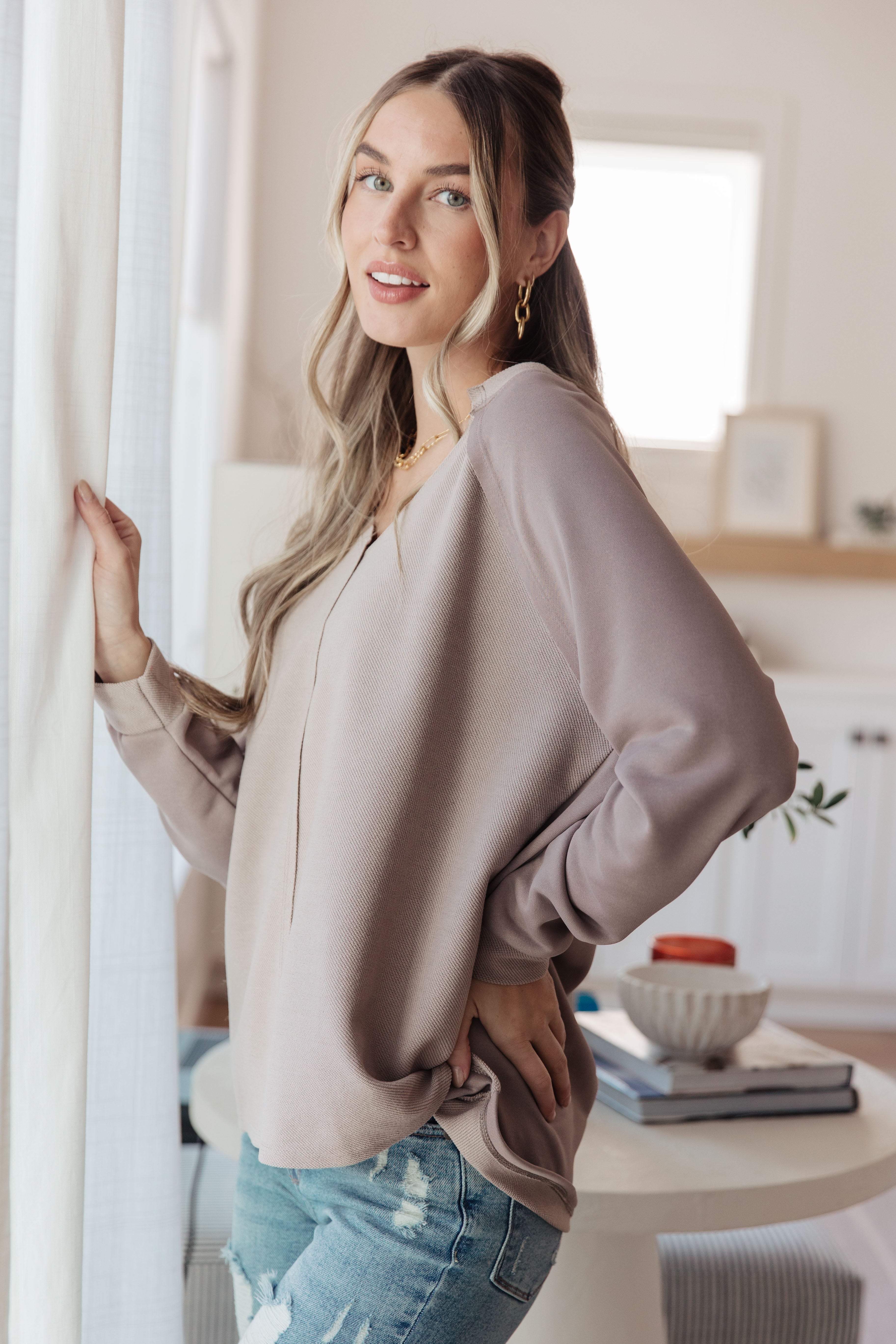 Woman wearing a loose beige V-neck top with long sleeves paired with distressed light blue jeans standing indoors by a curtain