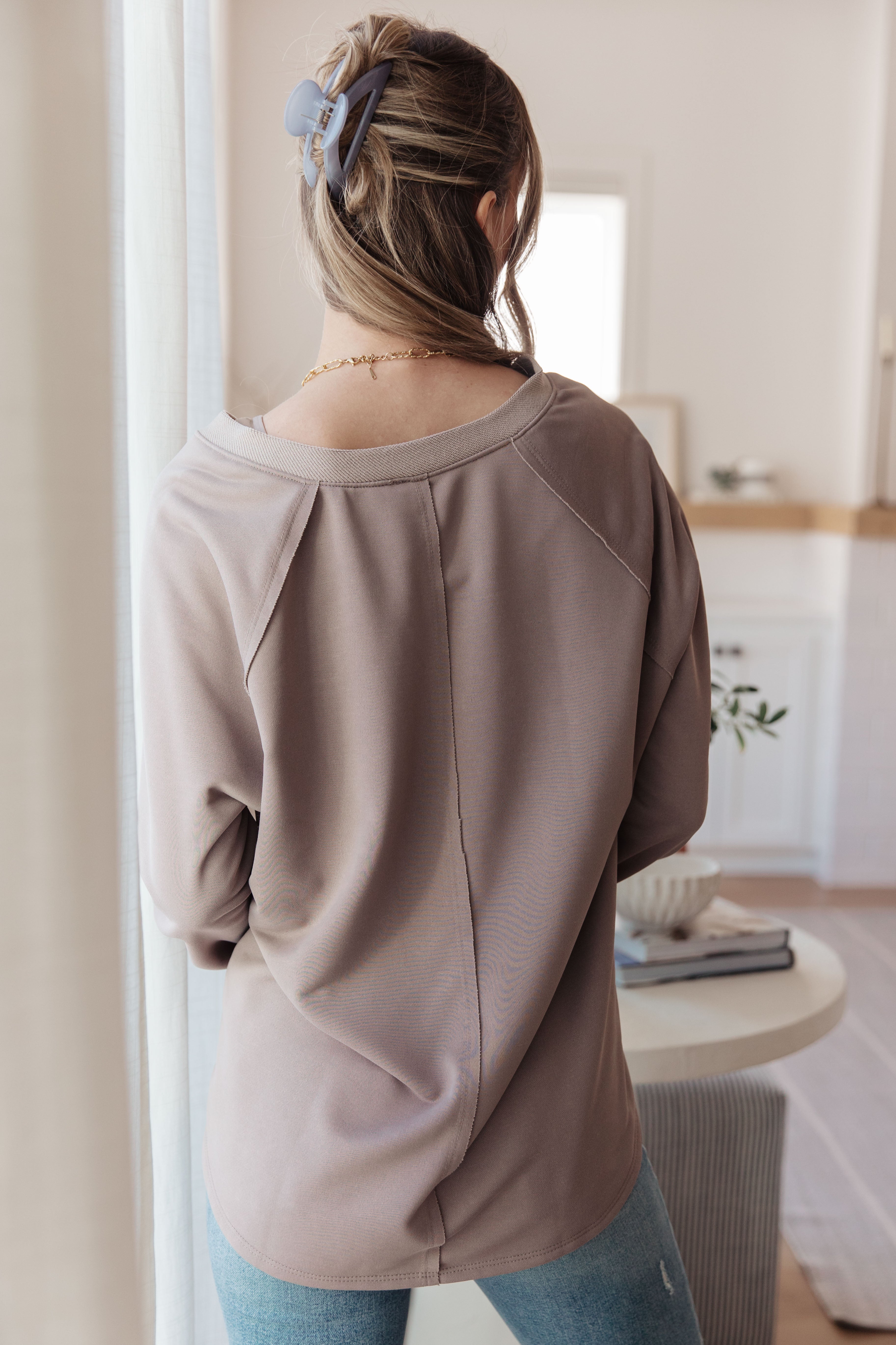 Woman wearing a beige loose long sleeve top with seam details on the back standing indoors by a window