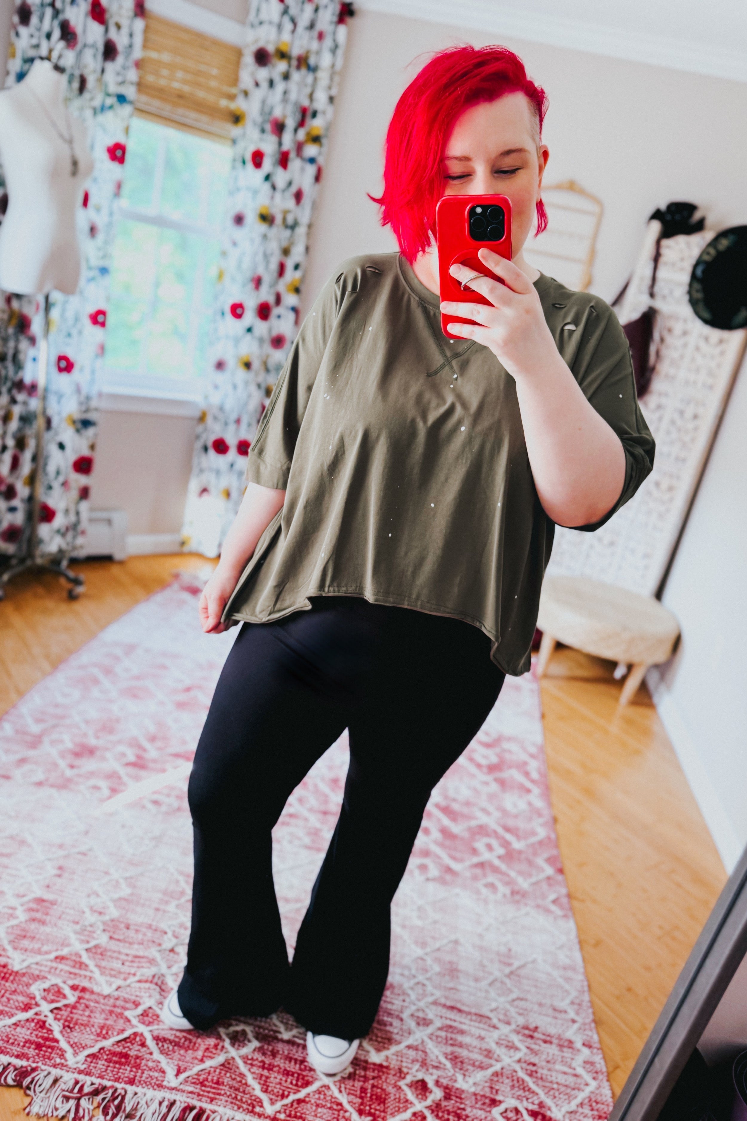Woman taking mirror selfie wearing an olive loose fit short sleeve top with subtle white speckled spots and black pants indoors