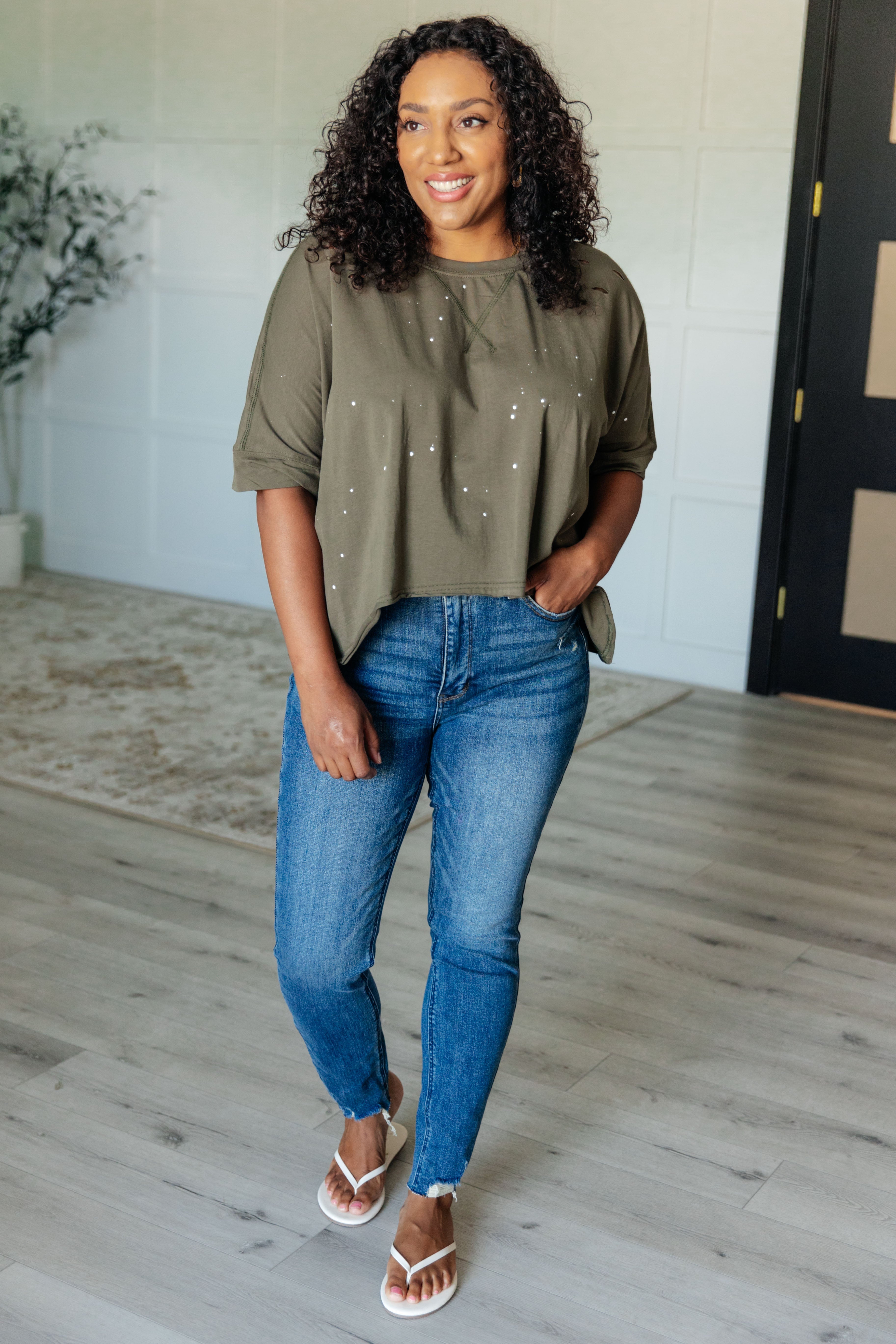Smiling woman indoors wearing an olive relaxed fit short sleeve top with distressed details and blue jeans