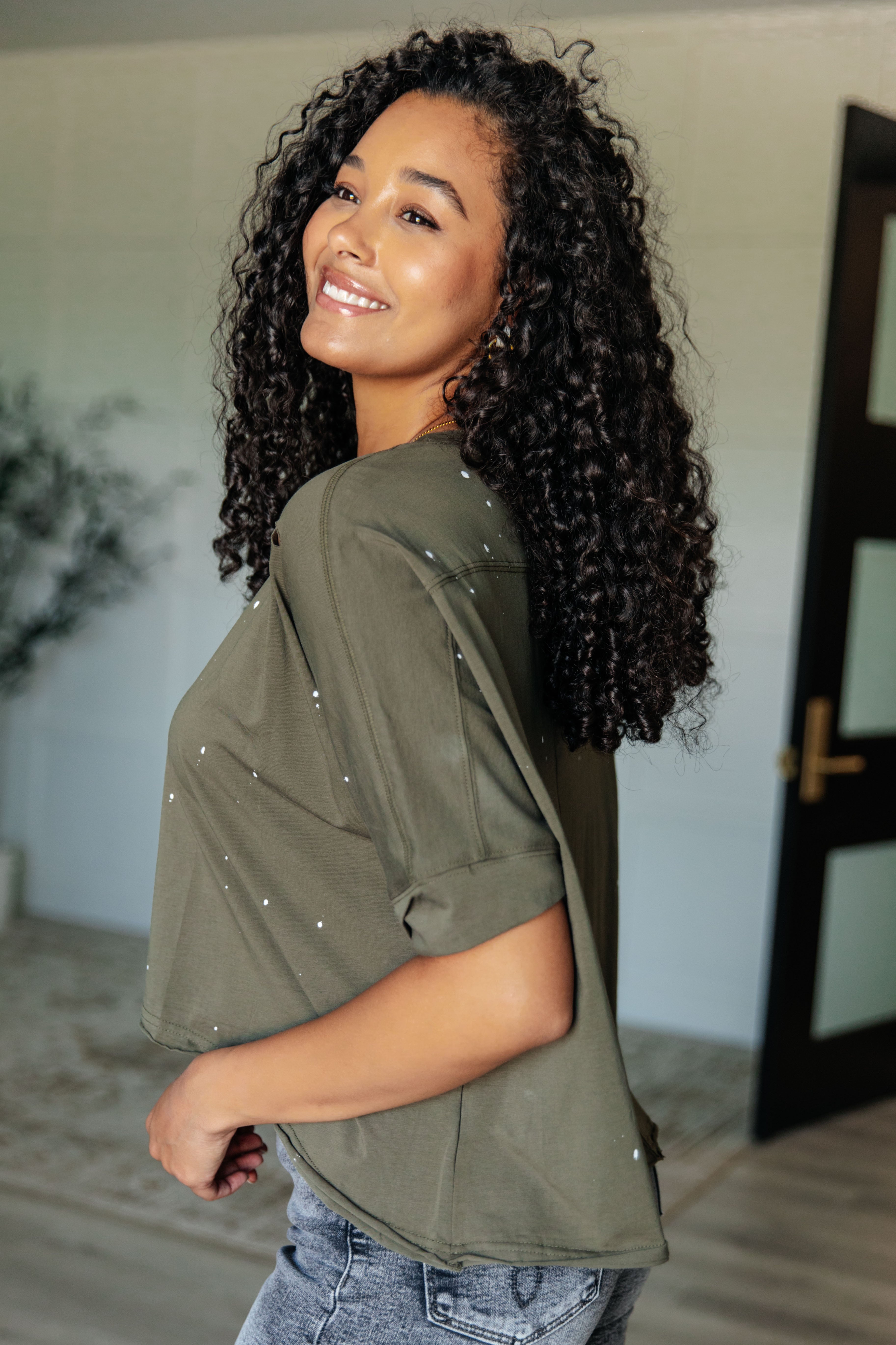 Smiling woman indoors wearing an olive loose fit short sleeve top with subtle distressing and light wash jeans