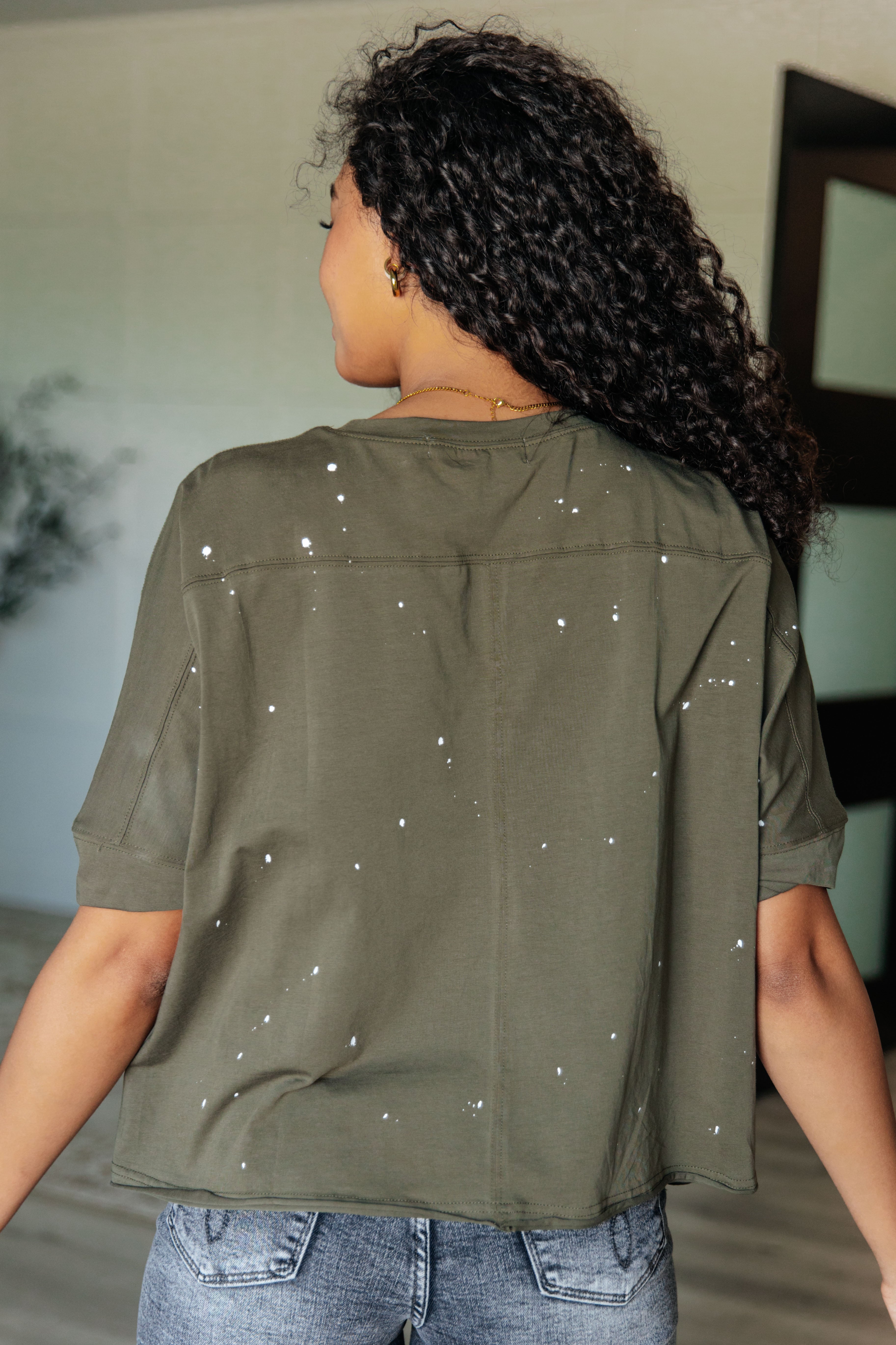 Woman seen from behind wearing an olive relaxed fit short sleeve top with white paint splatter paired with light wash jeans indoors