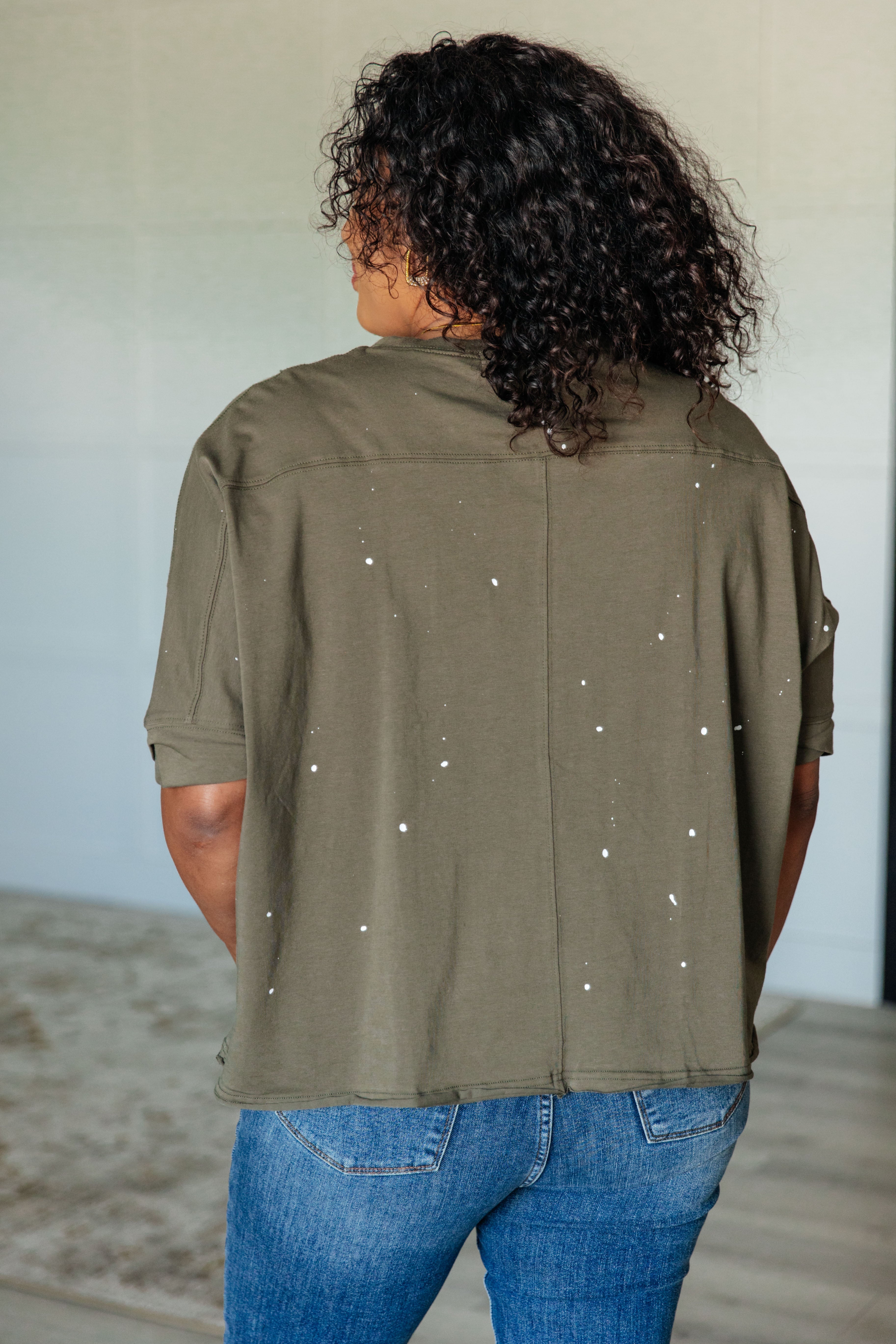 Woman with curly hair seen from behind wearing an olive relaxed fit short sleeve top with white paint splatter and blue jeans indoors