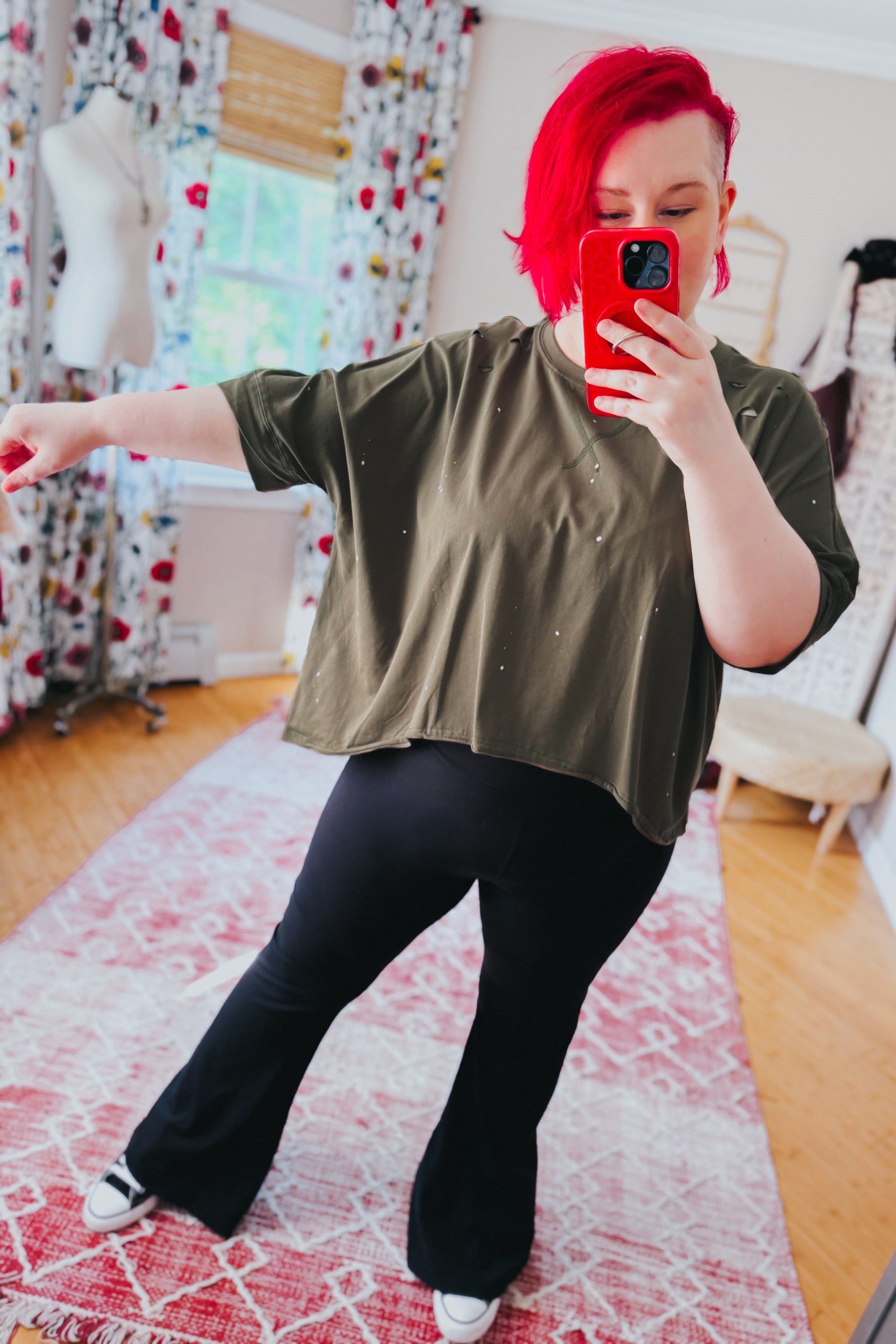 Woman with vibrant red hair taking selfie wearing olive loose fit short sleeve top with paint speckle and black flared pants indoors
