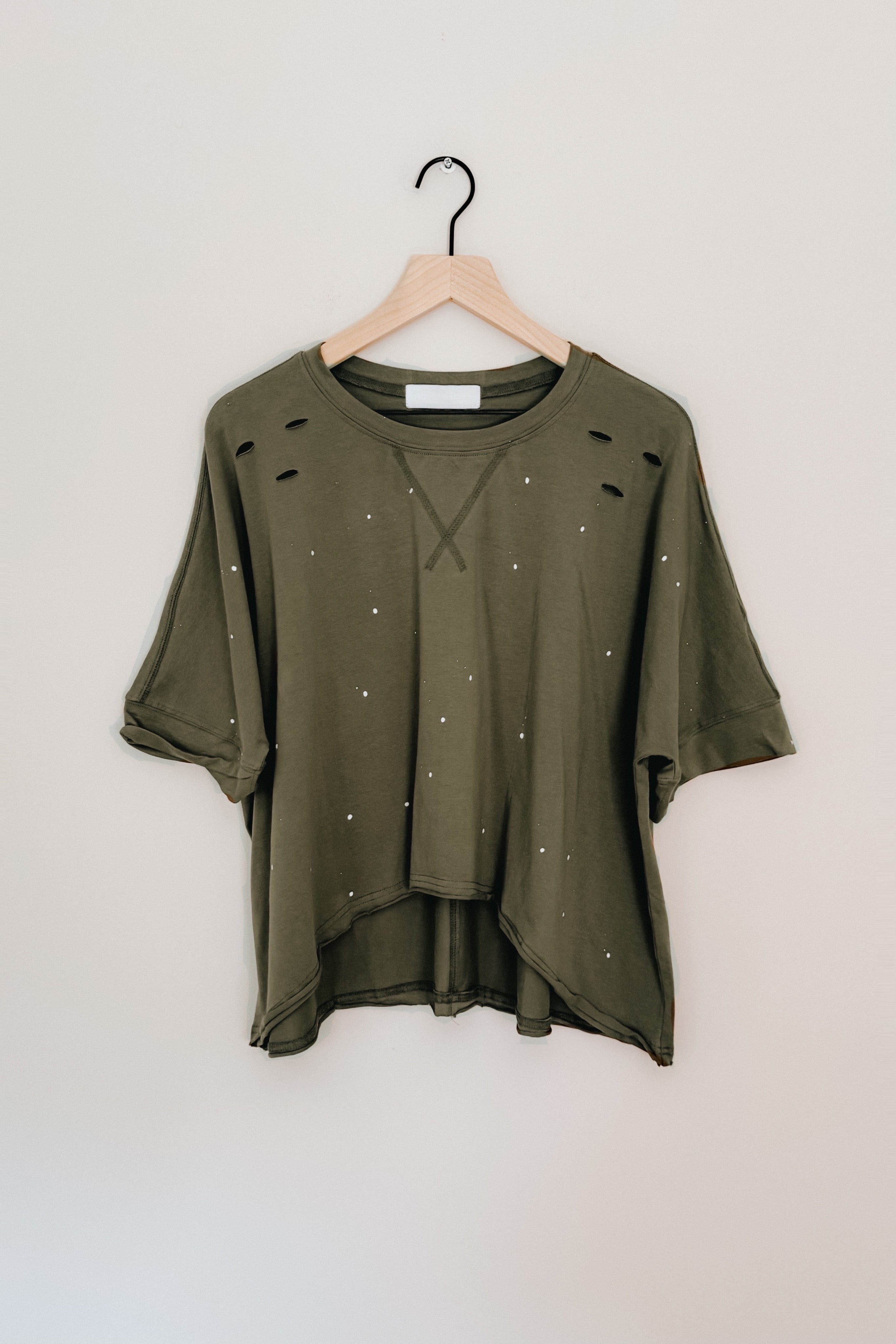 Olive oversized short sleeve top with distressed holes and white speckled details hanging on wooden hanger