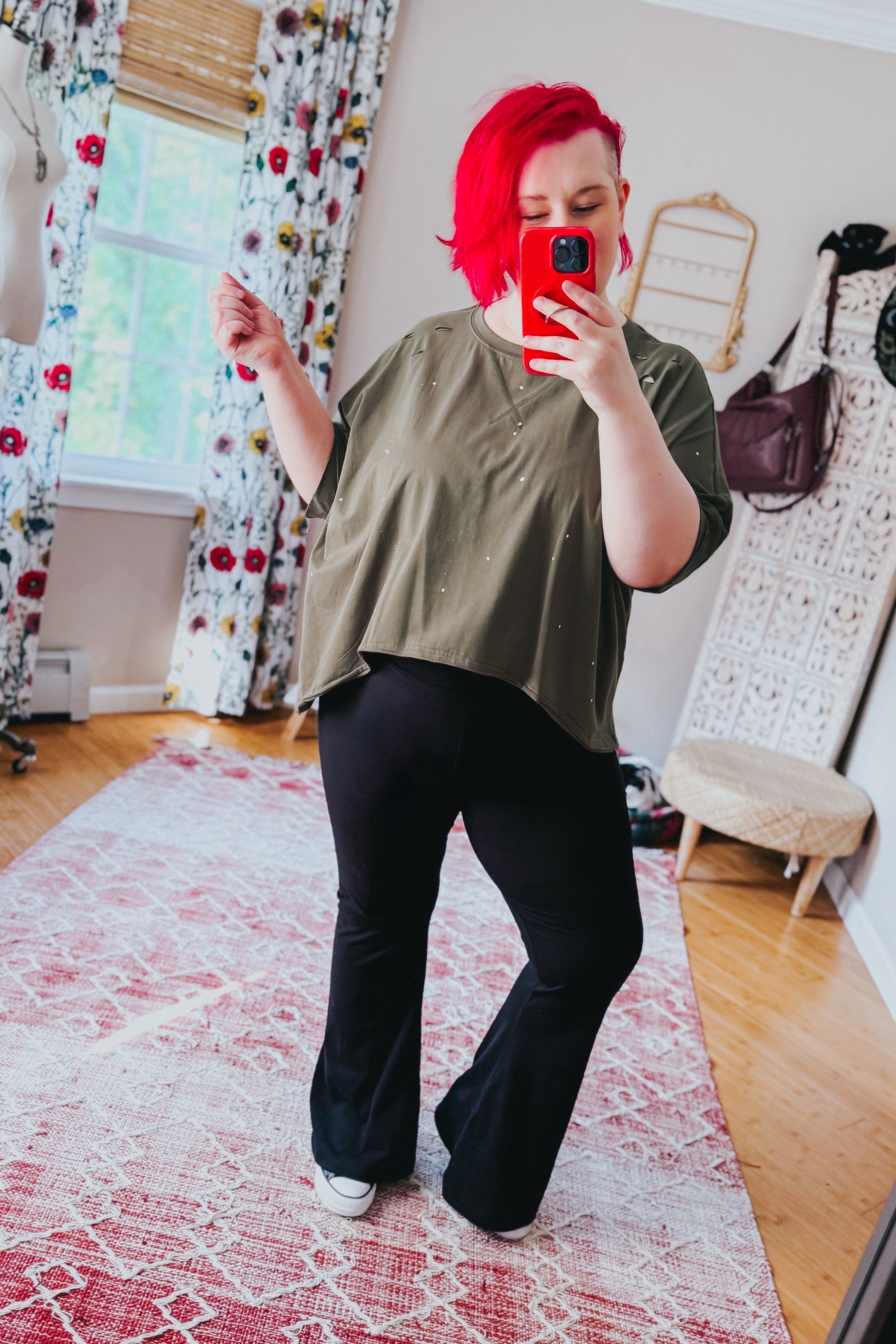 Woman with vibrant red hair taking mirror selfie wearing an olive oversized short sleeve top with small speckled spots and black flared pants indoors