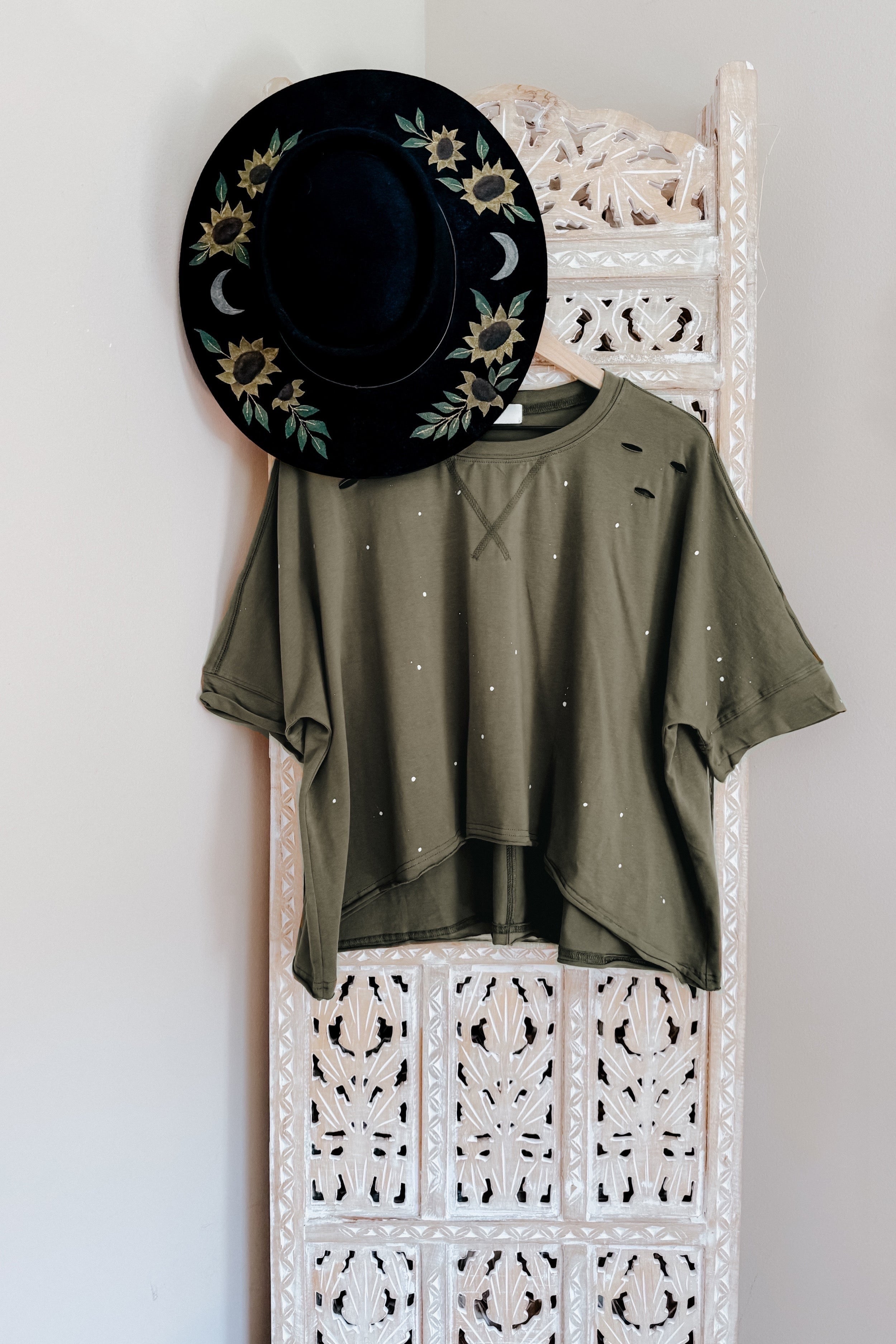 Olive relaxed fit short sleeve top with small white splatter and distressing paired with a black floral wide-brim hat hanging on carved wooden screen