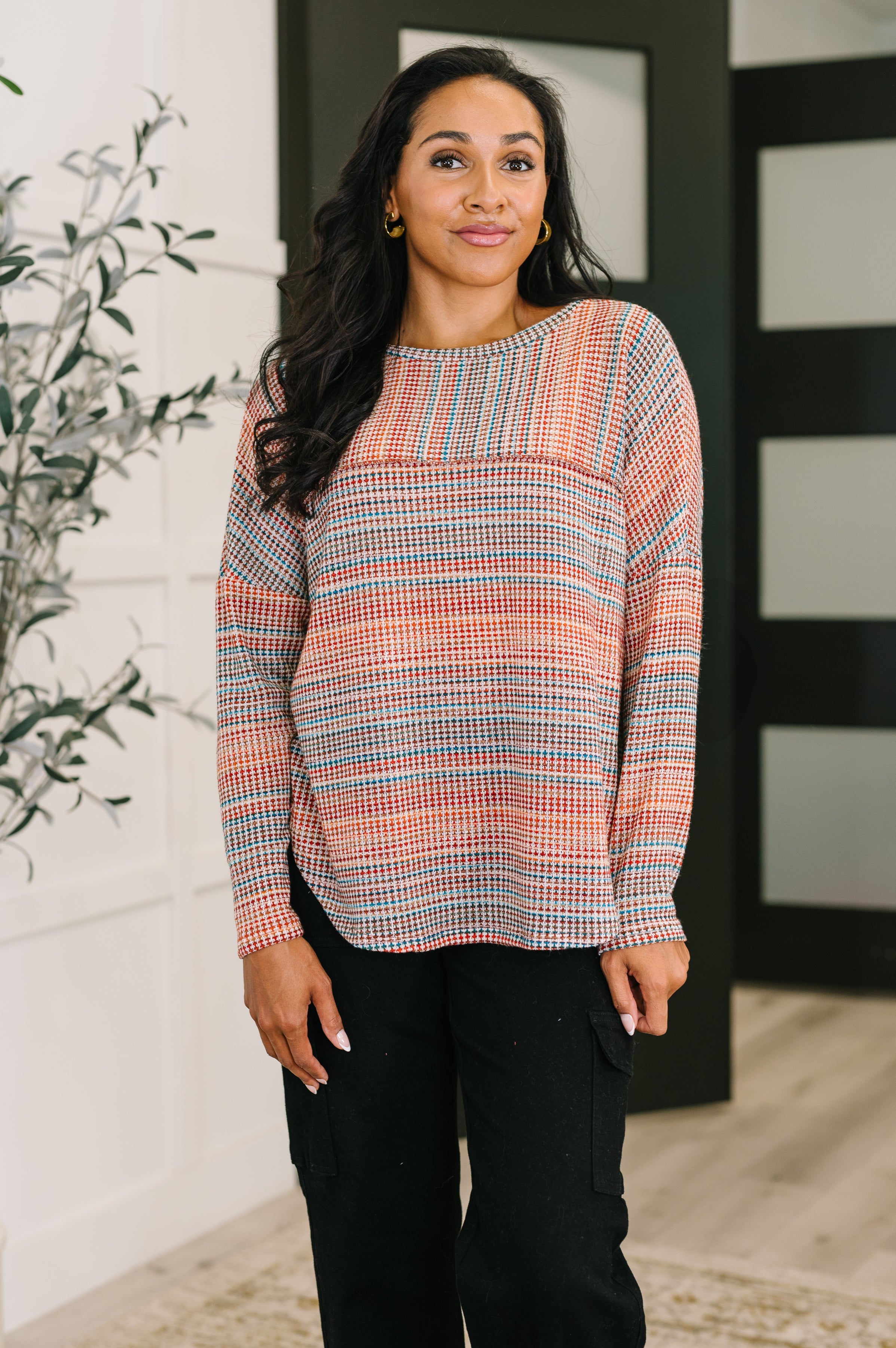Woman indoors wearing a long sleeve orange patterned top paired with black pants standing near a plant and doors