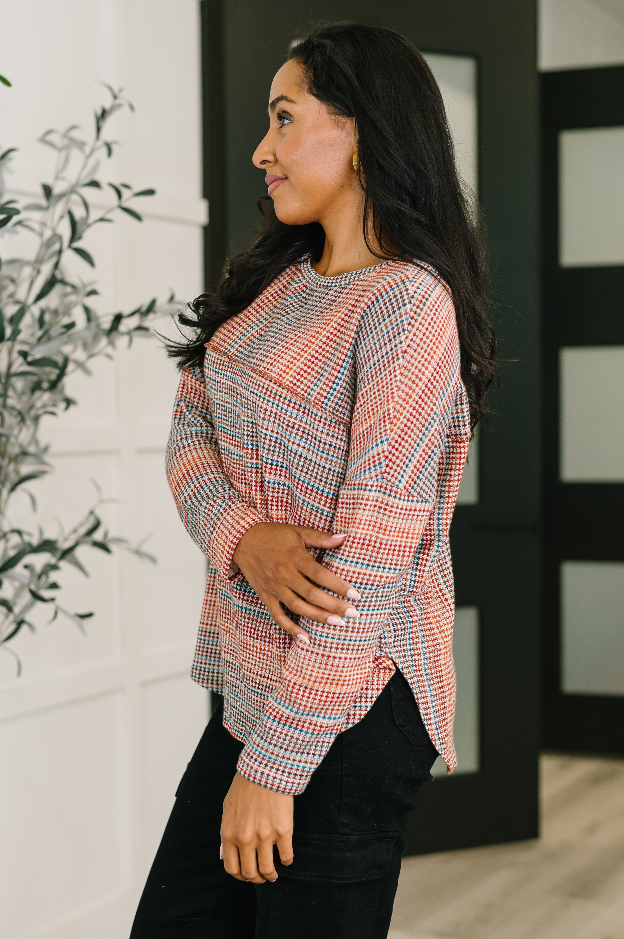 Woman wearing an orange textured long sleeve top with loose fit and black pants standing indoors near plant and doors