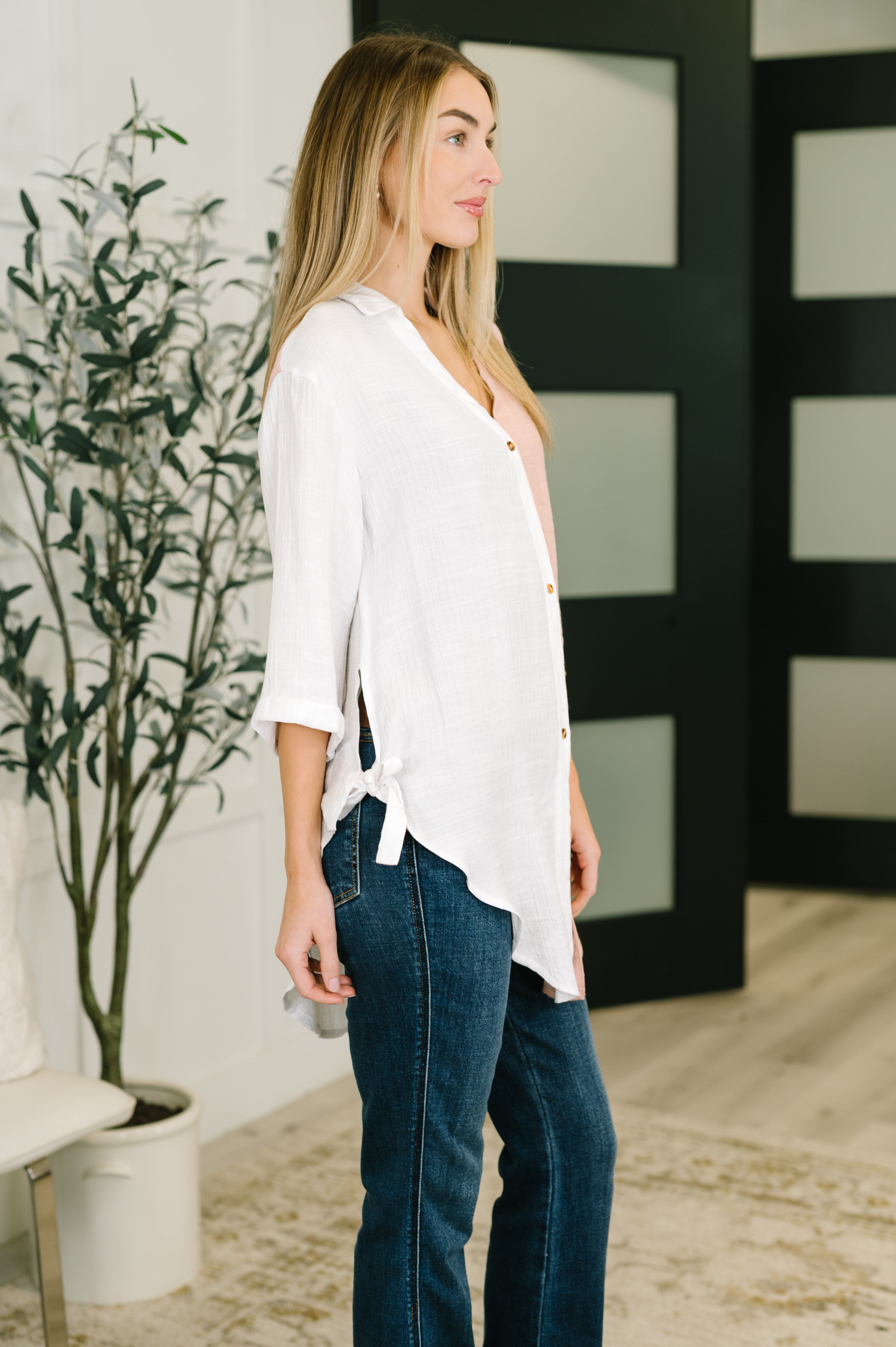 Woman wearing a white button-down top with side ties over a blush pink camisole and dark blue jeans indoors