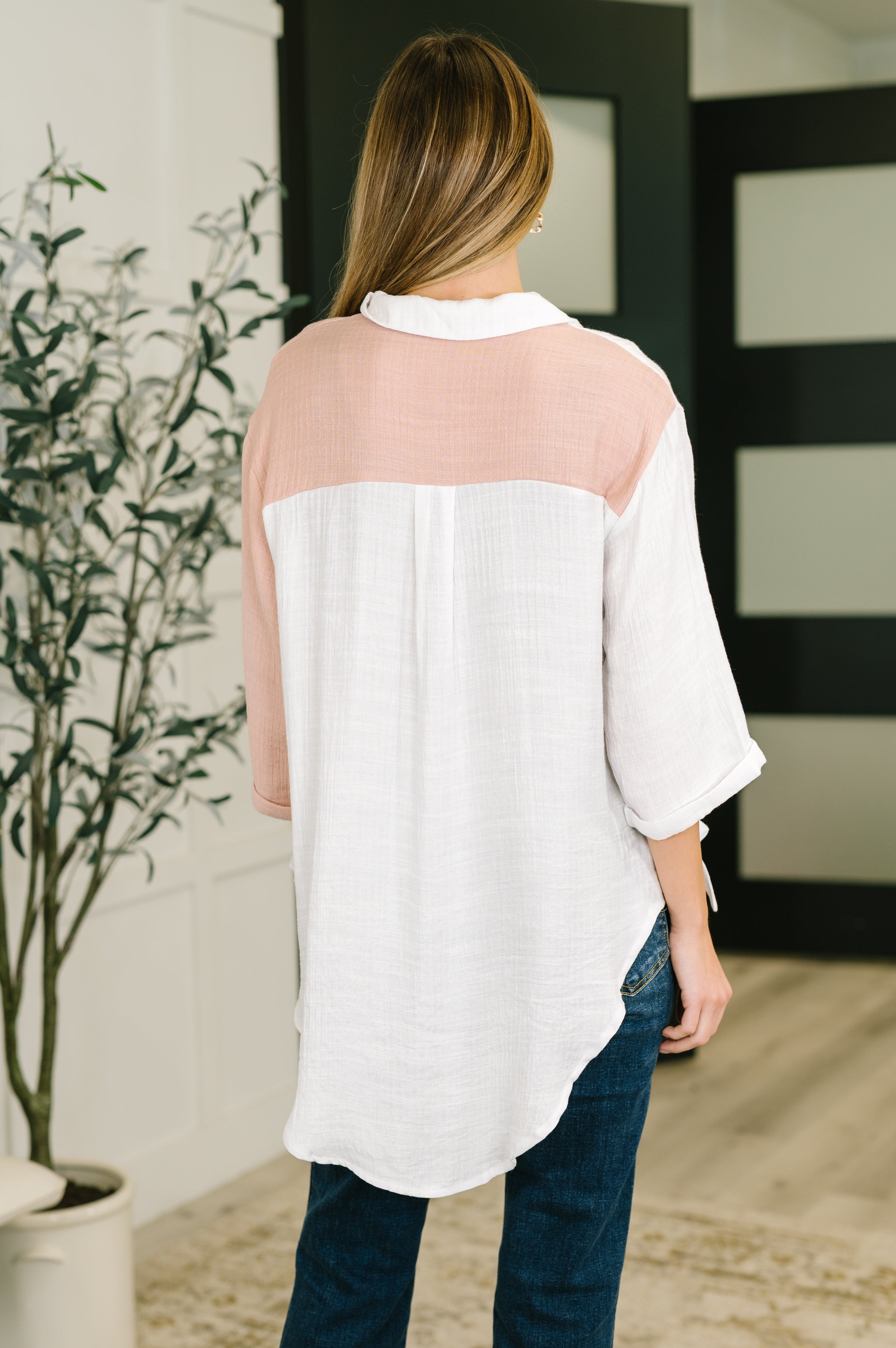 Woman wearing a white and blush pink dual toned button down top with rolled sleeves and dark blue jeans, standing indoors
