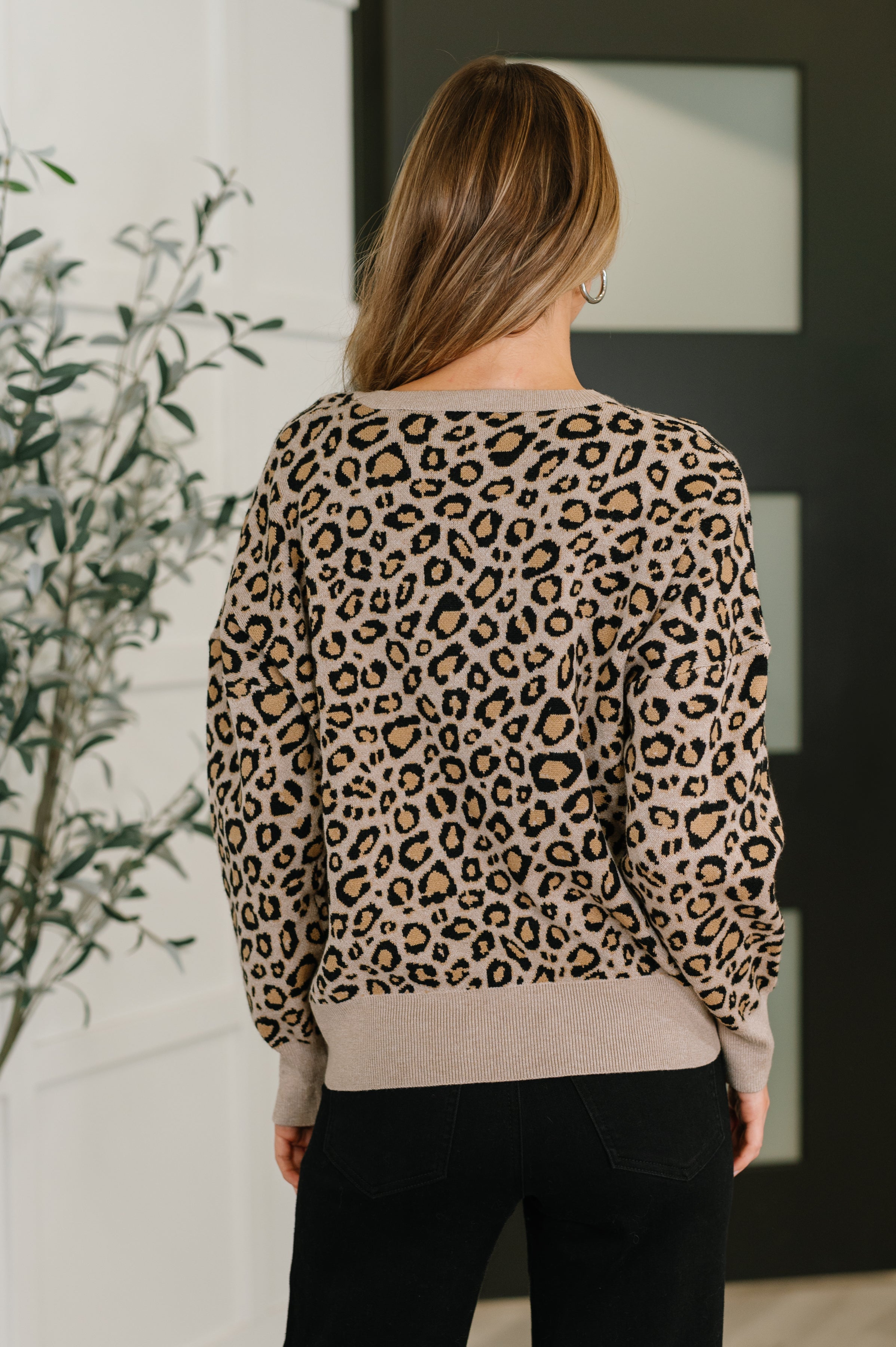 Woman shown from behind wearing a brown leopard print sweater paired with black pants indoors near a plant
