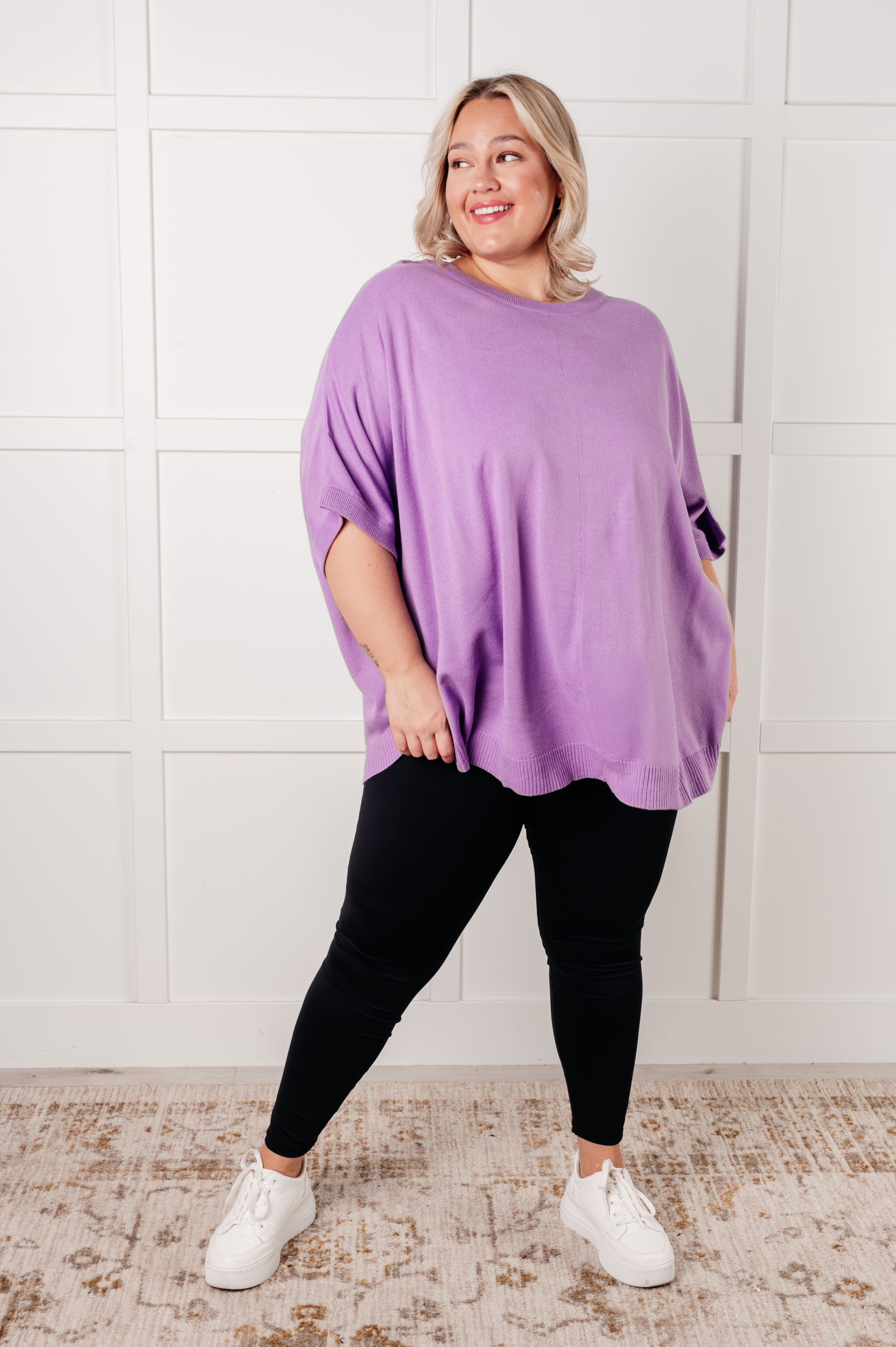 Plus size person wearing a loose lilac knit top with dolman sleeves paired with black leggings and white sneakers indoors