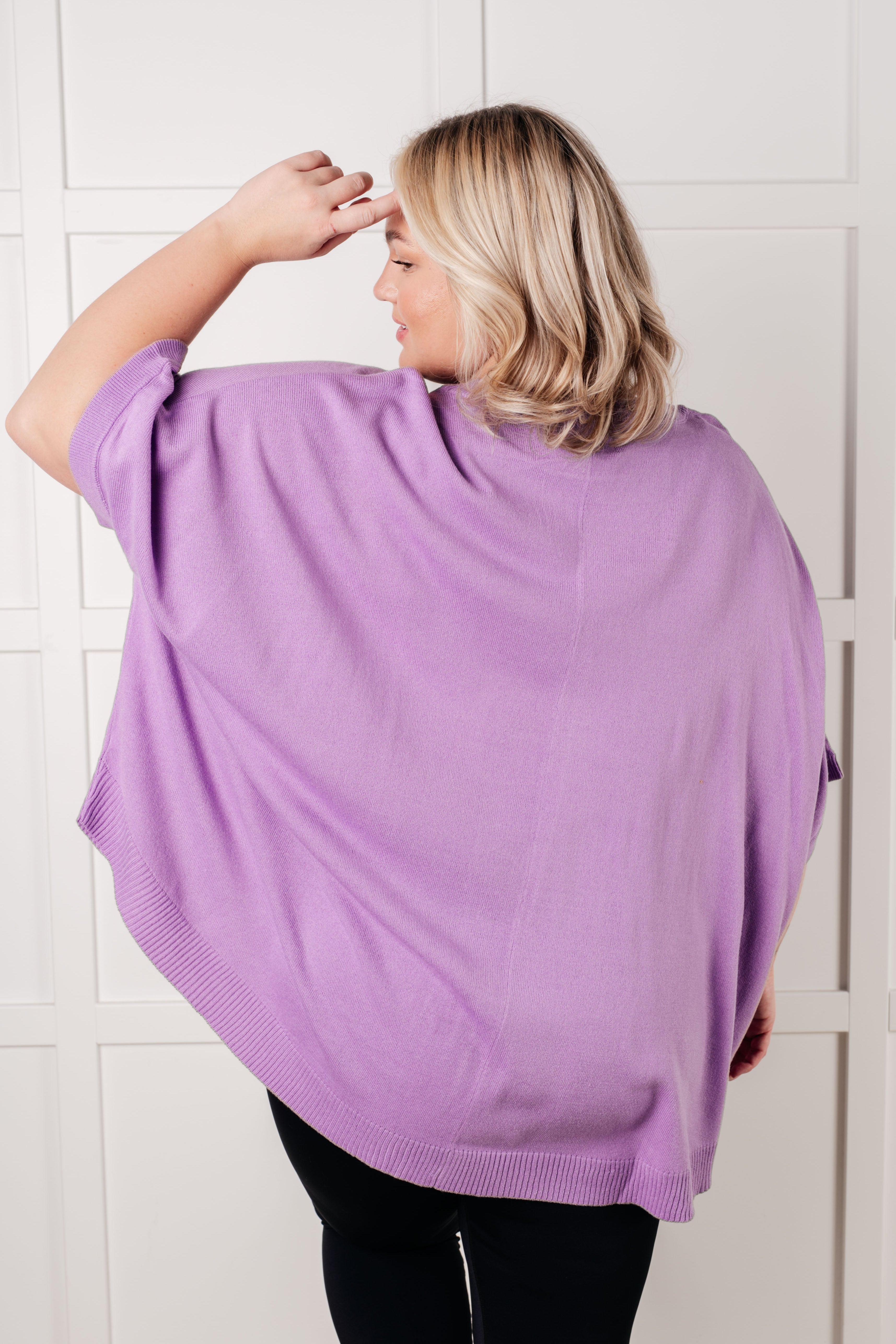 Woman seen from behind wearing a soft lilac dolman sleeve top with a relaxed fit and ribbed hem against a white wall