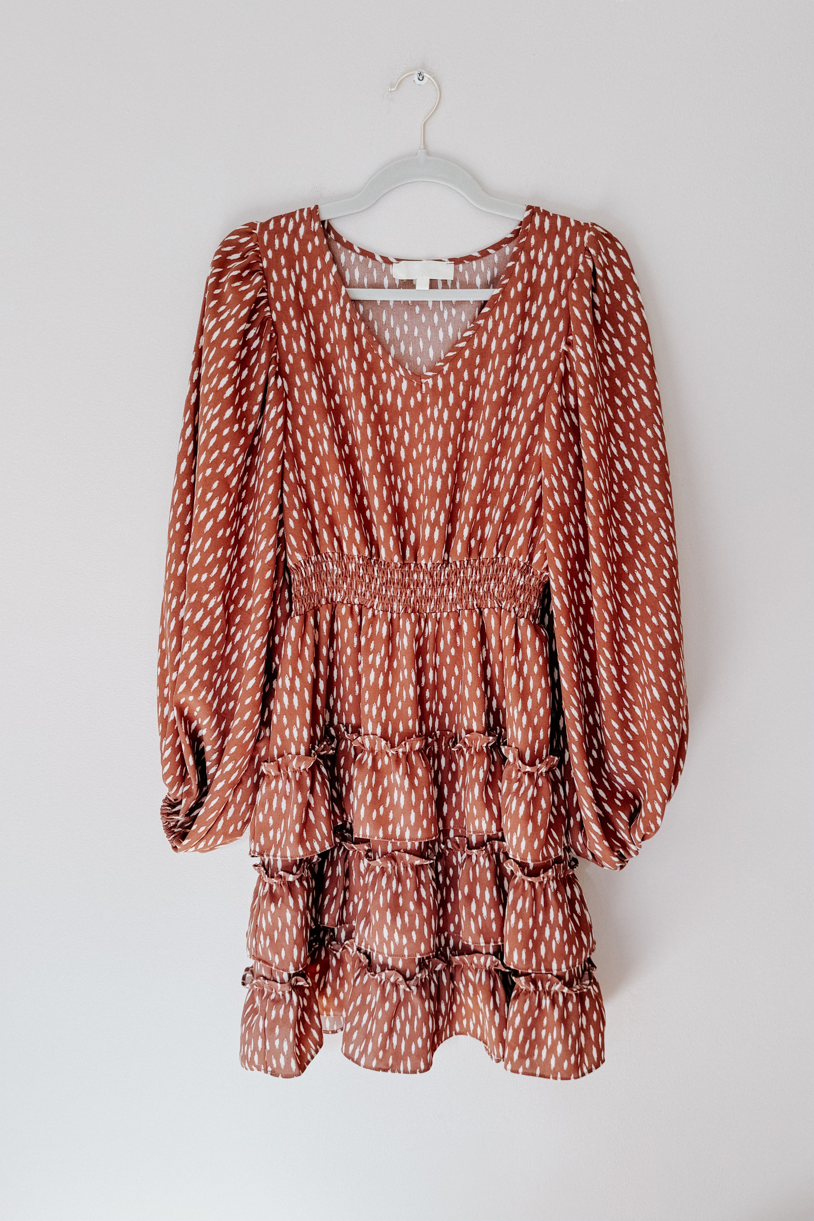 Terracotta dress featuring a white dash print, smocked waist, long voluminous sleeves, and tiered ruffled hem hanging on hanger