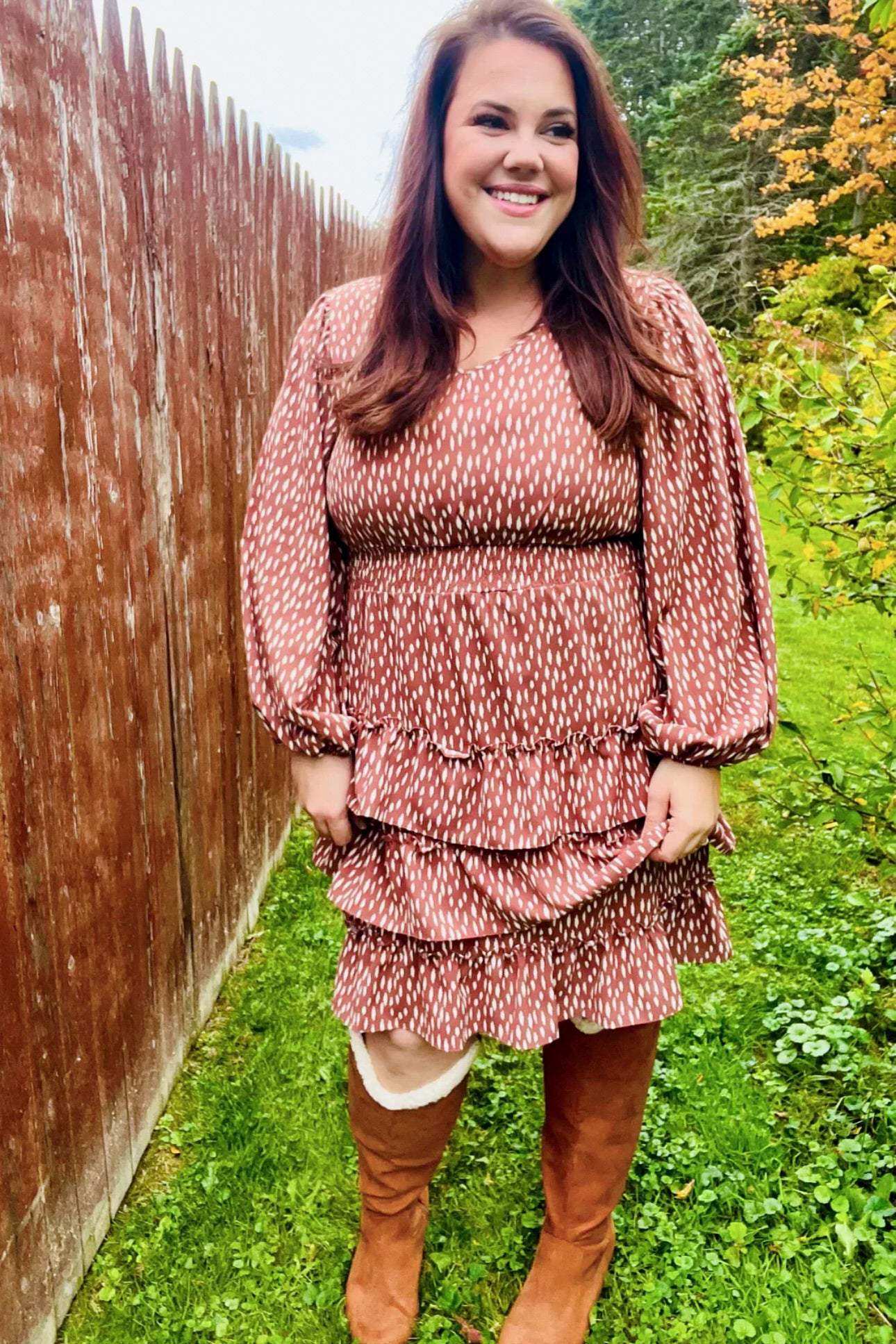 Woman smiling outdoors wearing a terracotta dress with abstract white spots, puffed sleeves, ruffled tiers, and brown knee-high boots