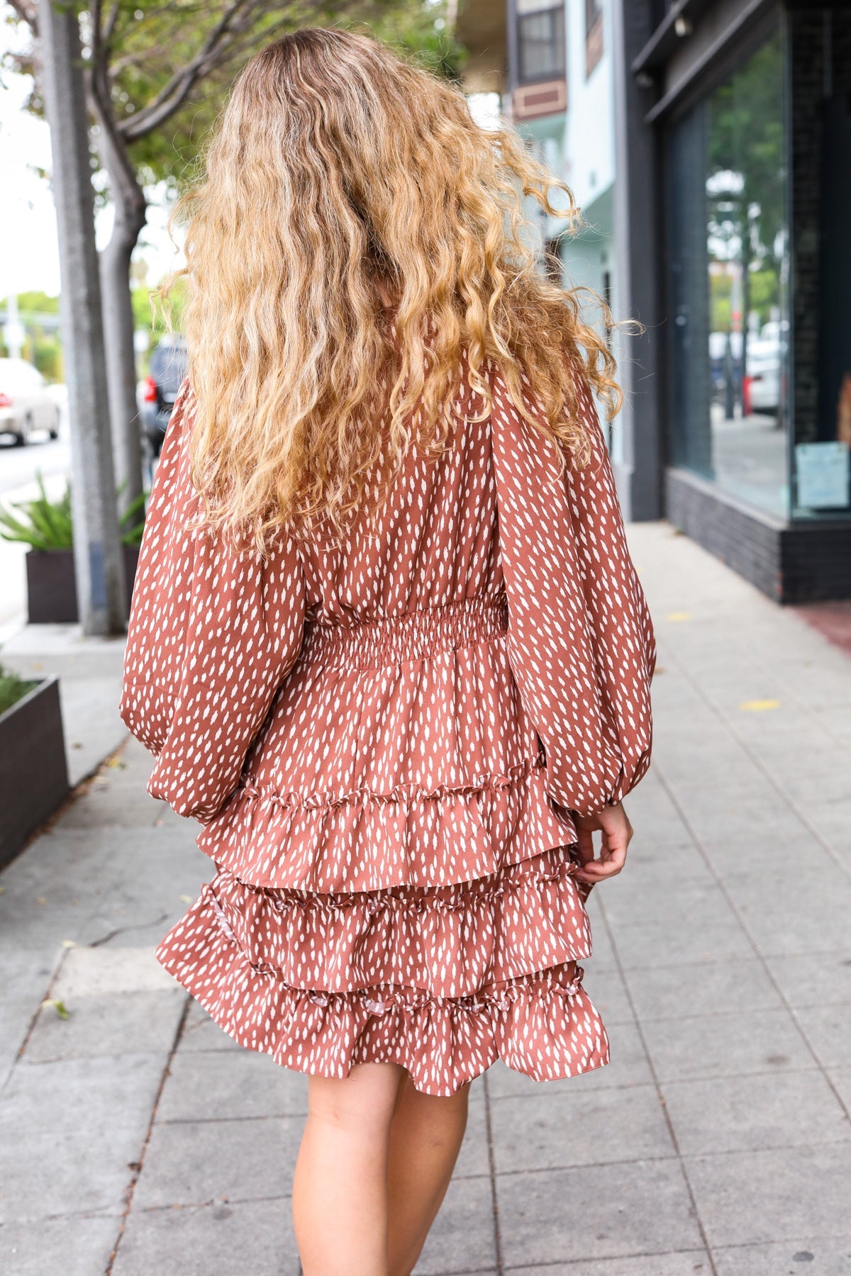 Woman walking outdoors wearing a terracotta dress with white abstract print and tiered ruffle skirt viewed from behind