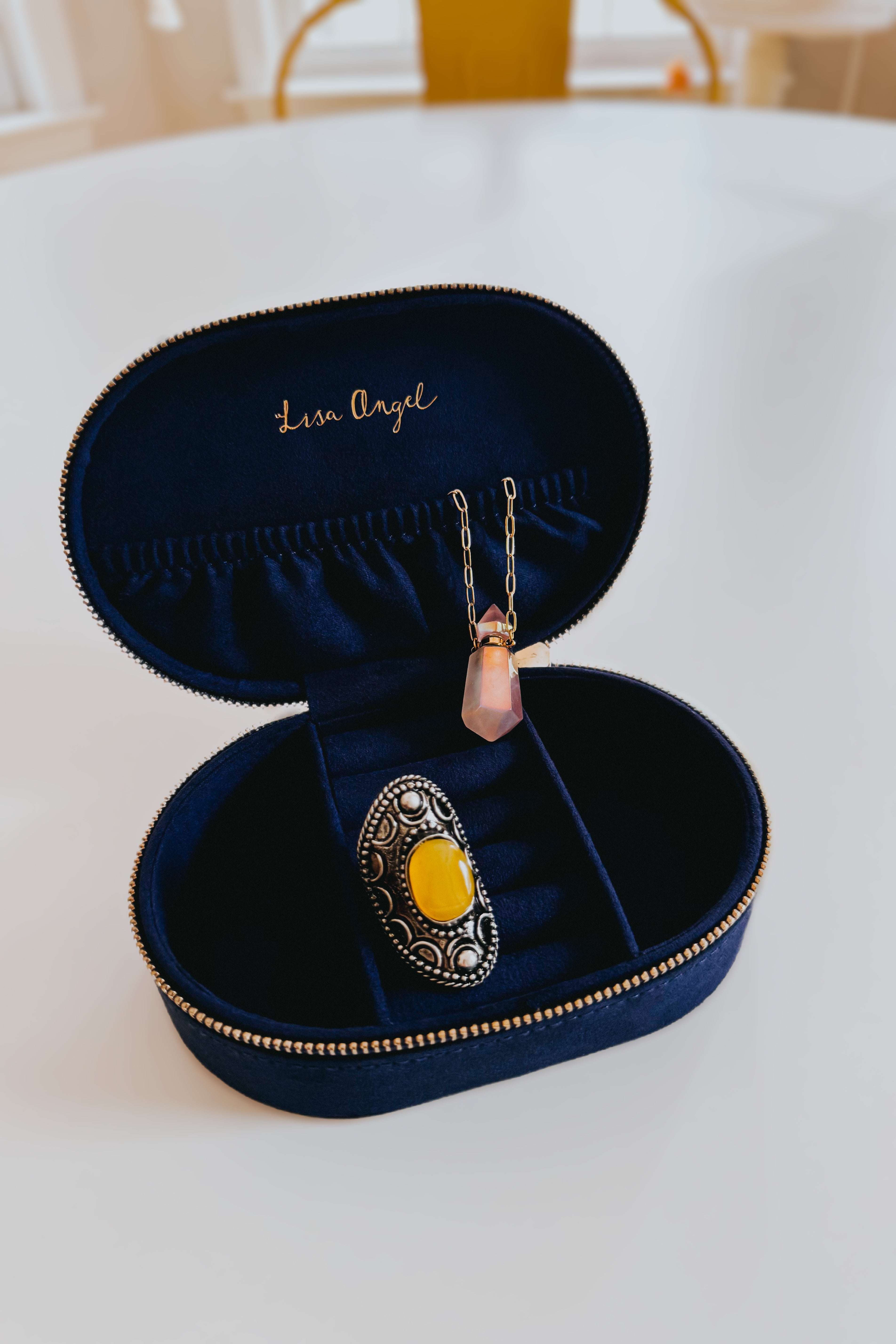 Oval navy velvet jewelry case with gold zipper containing yellow stone ring and delicate pendant necklace