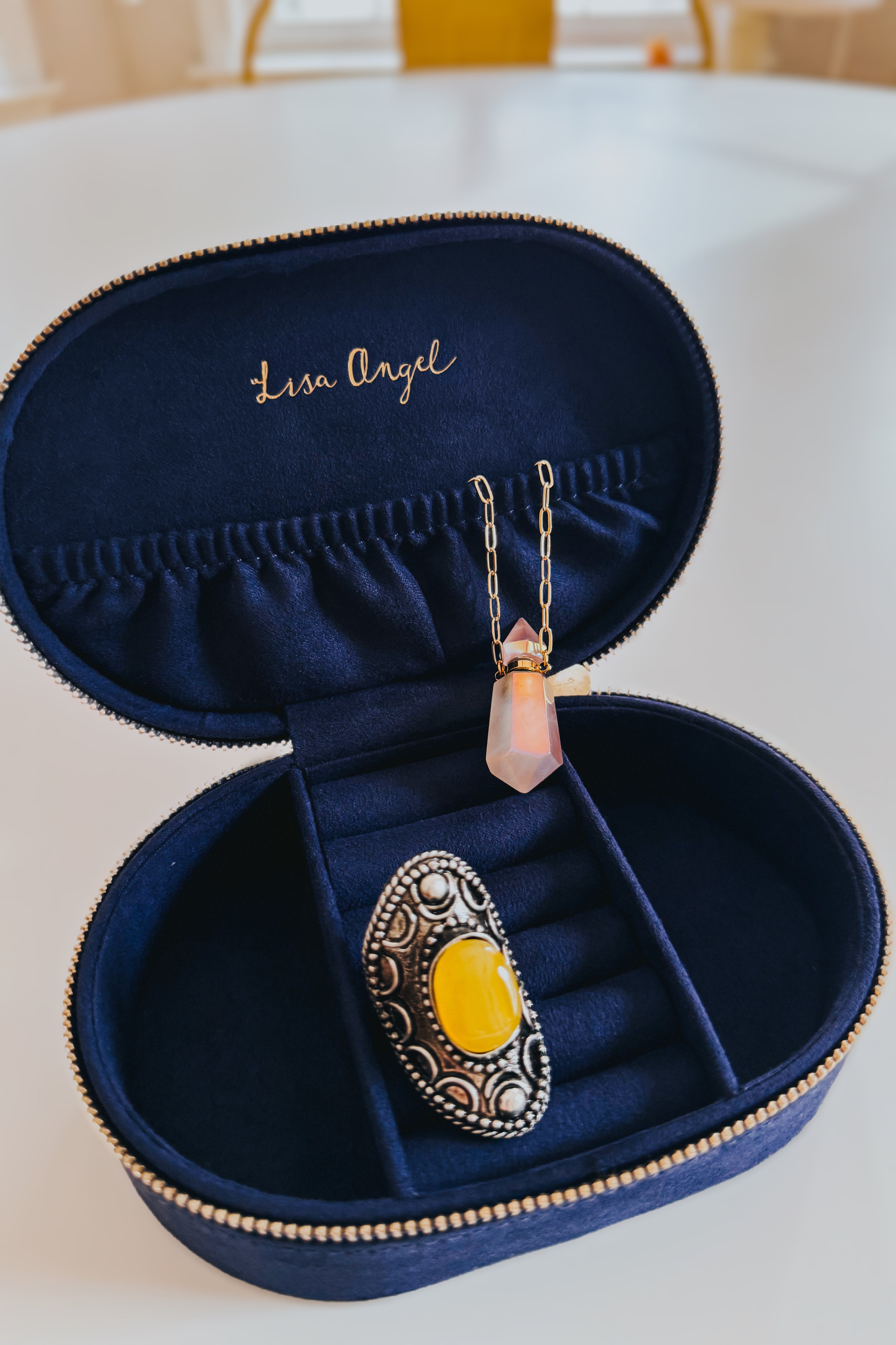 Navy velvet jewelry case with gold zipper holding an ornate silver ring with yellow stone and a delicate pendant necklace