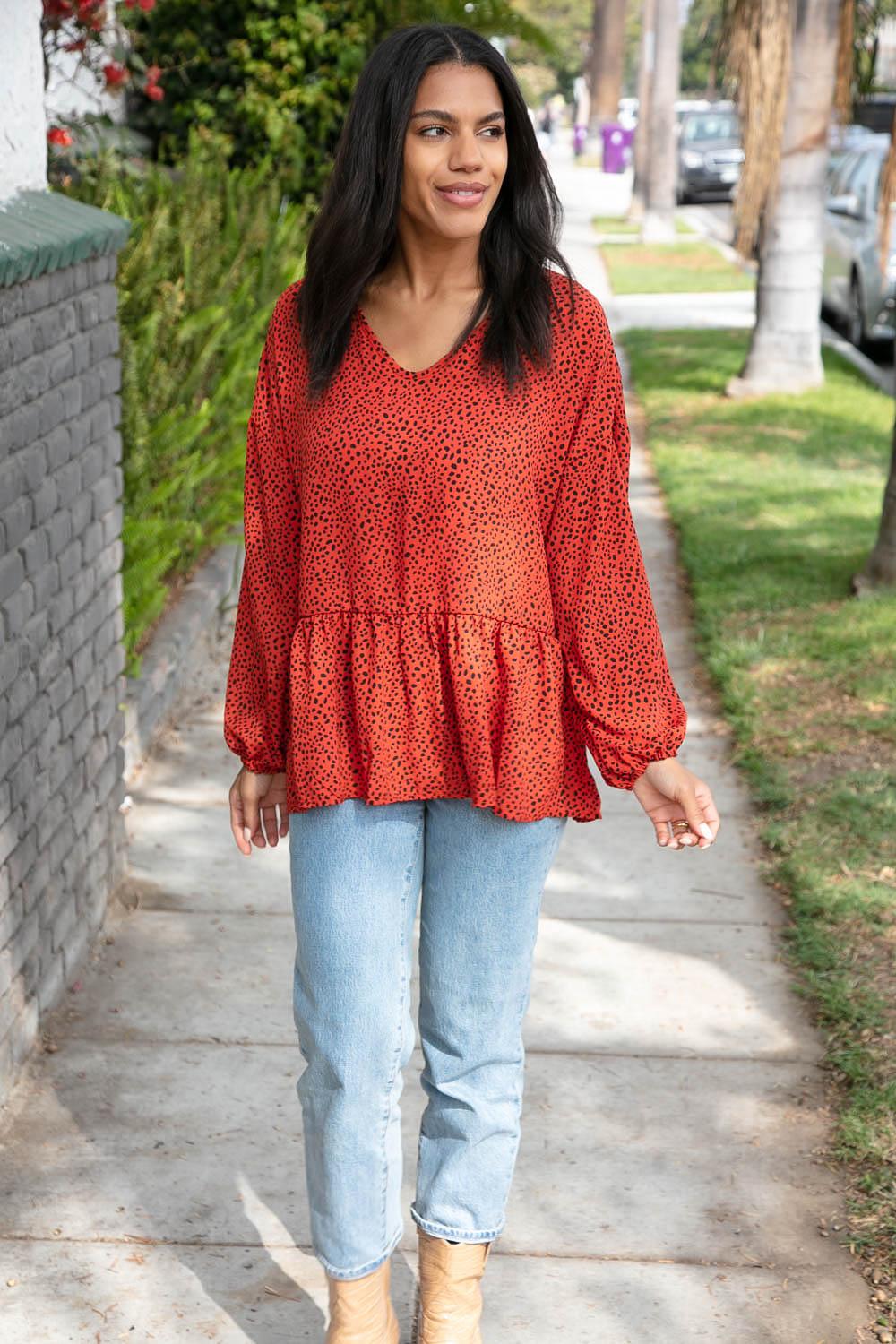 Woman walking on sidewalk wearing rust long-sleeve blouse with black dots and light blue jeans
