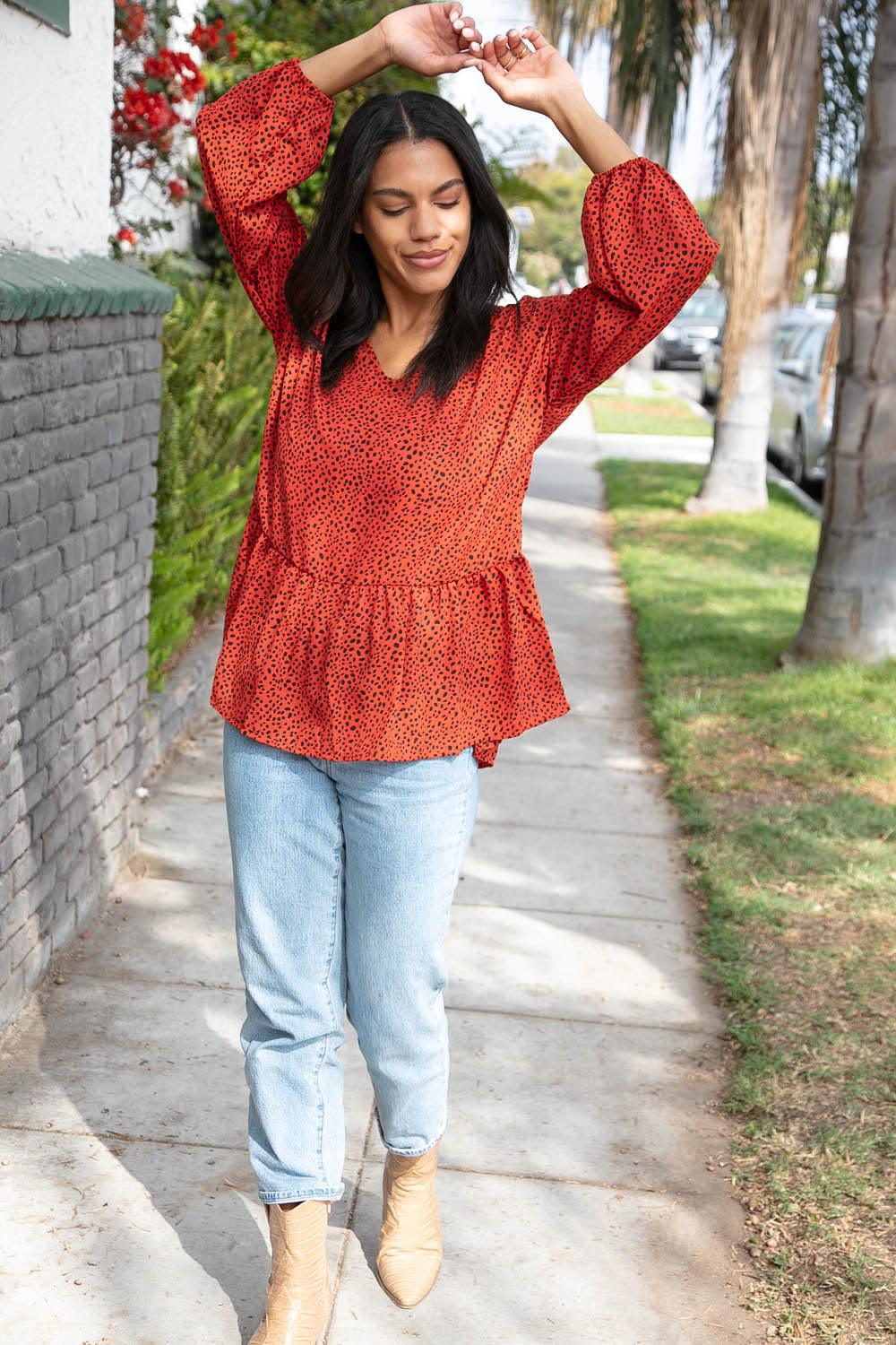 Woman walking on sidewalk wearing rust-colored blouse with black speckled print and light blue jeans