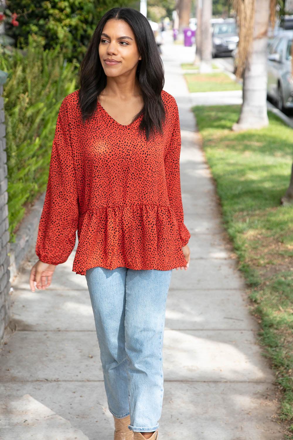 Woman walking along sidewalk wearing rust-colored blouse with small black dots and loose-fitting light blue jeans