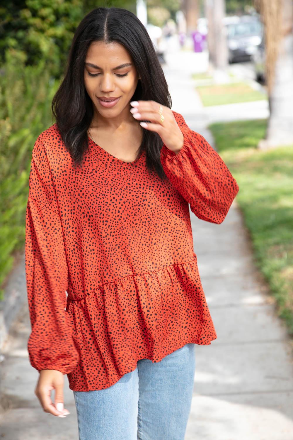 Woman walking outdoors wearing a rust blouse with small black spots and a gathered peplum hem paired with light blue jeans