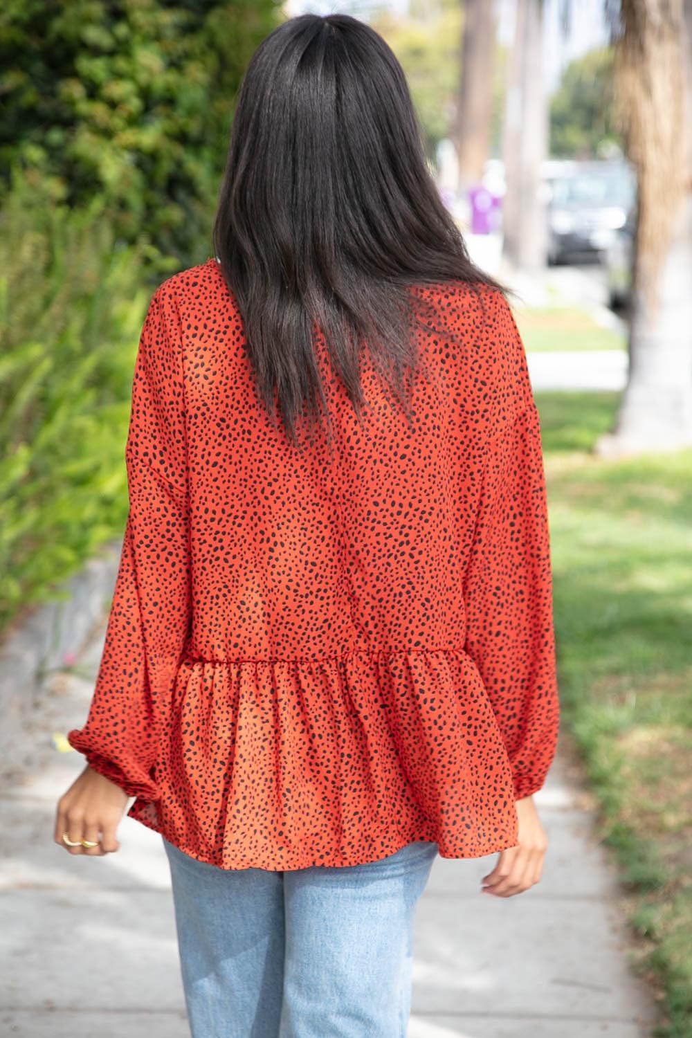 Woman seen from behind wearing a rust blouse with black speckles and a gathered ruffle hem paired with jeans outdoors
