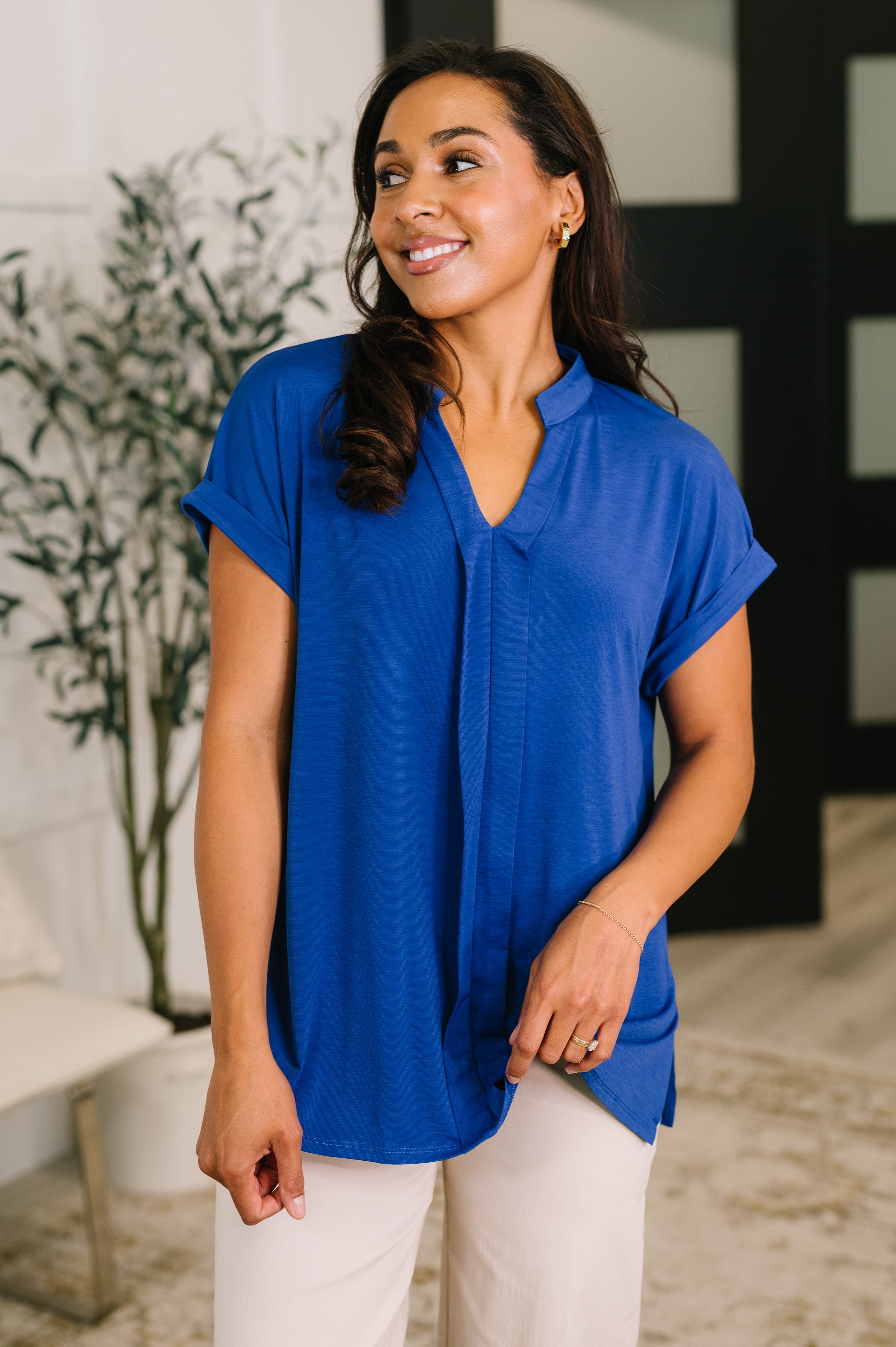 Woman wearing relaxed royal blue split neck blouse with rolled short sleeves indoors by plant and chair