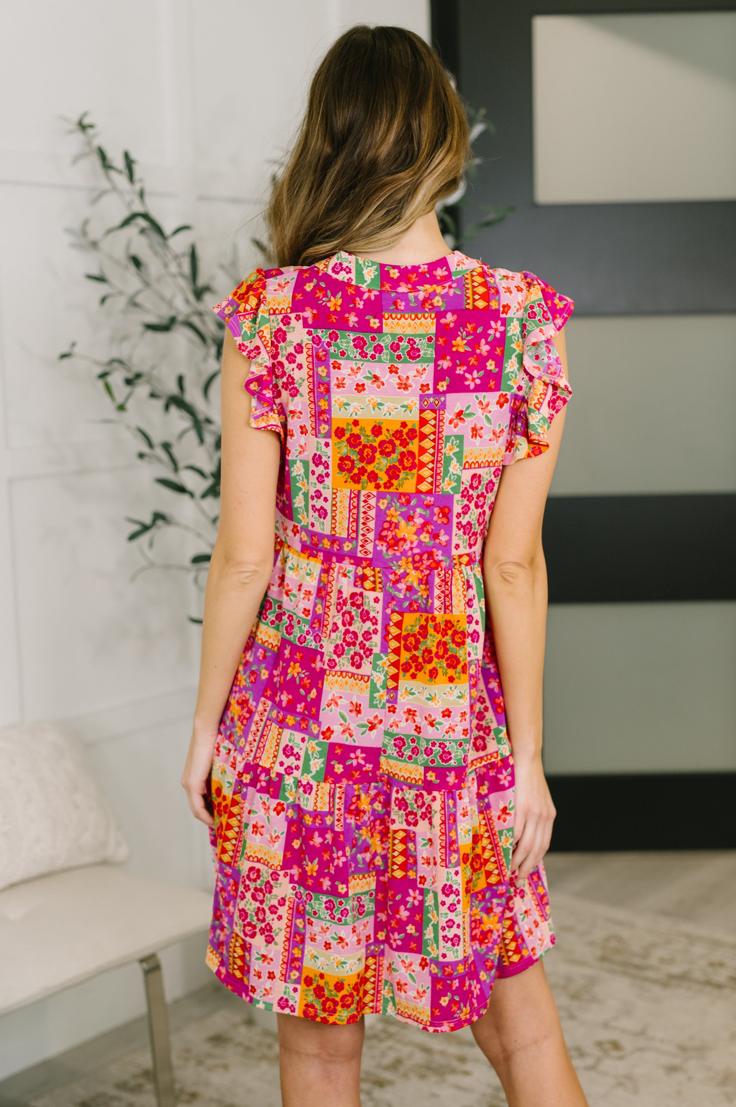 Back view of woman wearing fuchsia patchwork floral dress with flutter sleeves and gathered tiers indoors