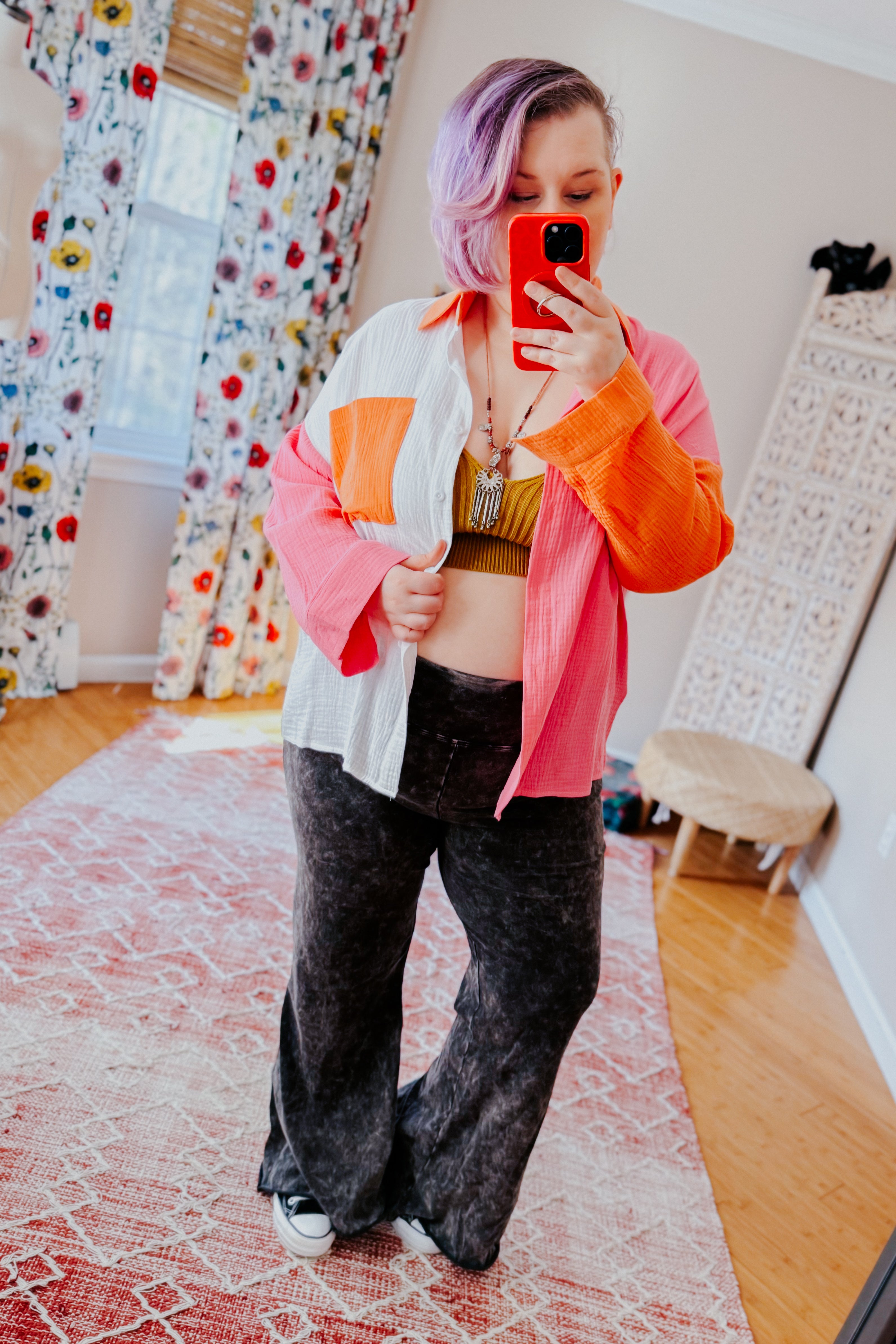 Woman with lavender hair wearing pink, orange, and ivory colorblock shirt over mustard crop top and black flared pants taking mirror selfie