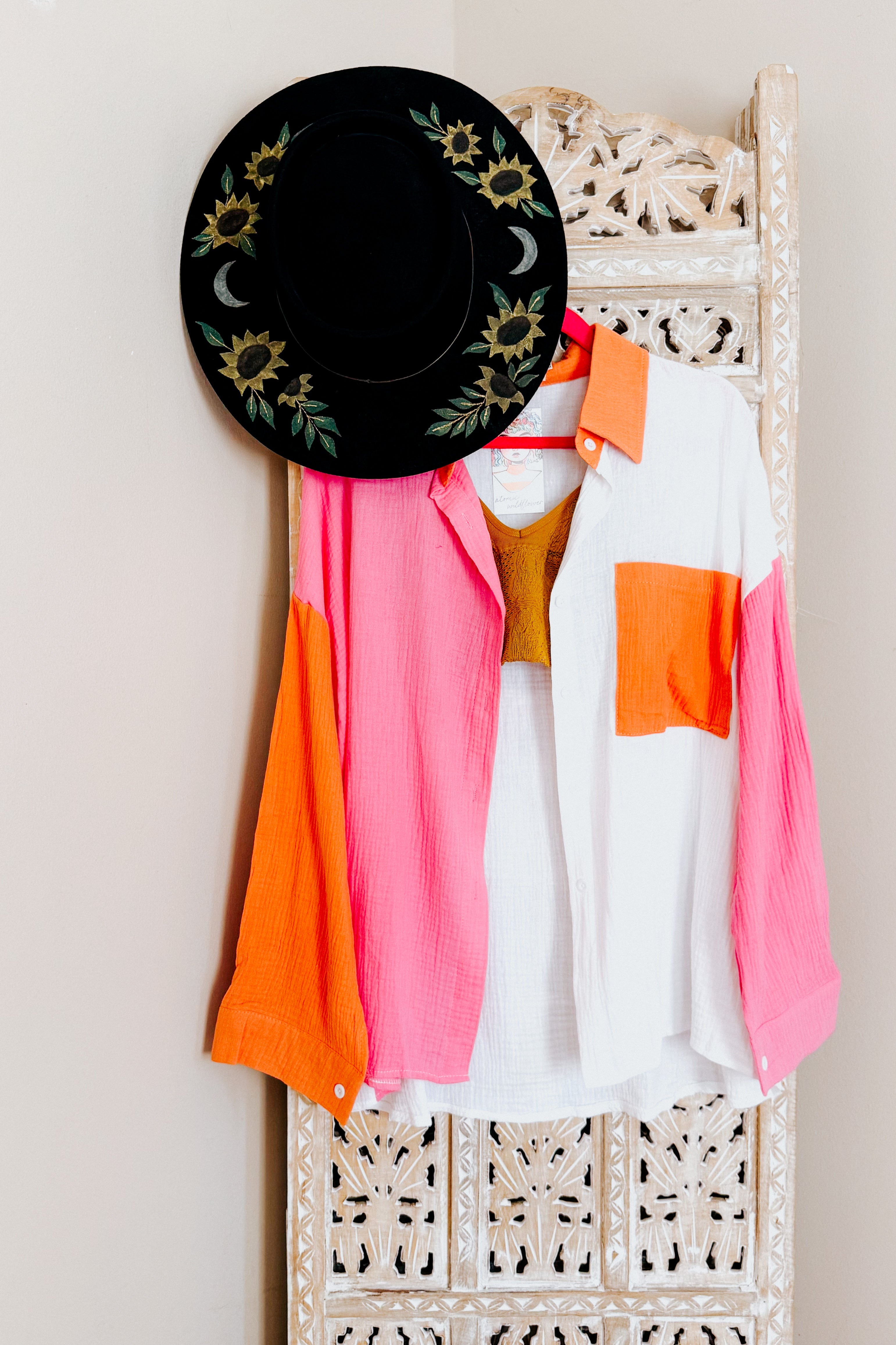 Black wide-brim hat with embroidered sunflowers and moons hanging above pink, orange, and ivory colorblock shirt on carved wood panel