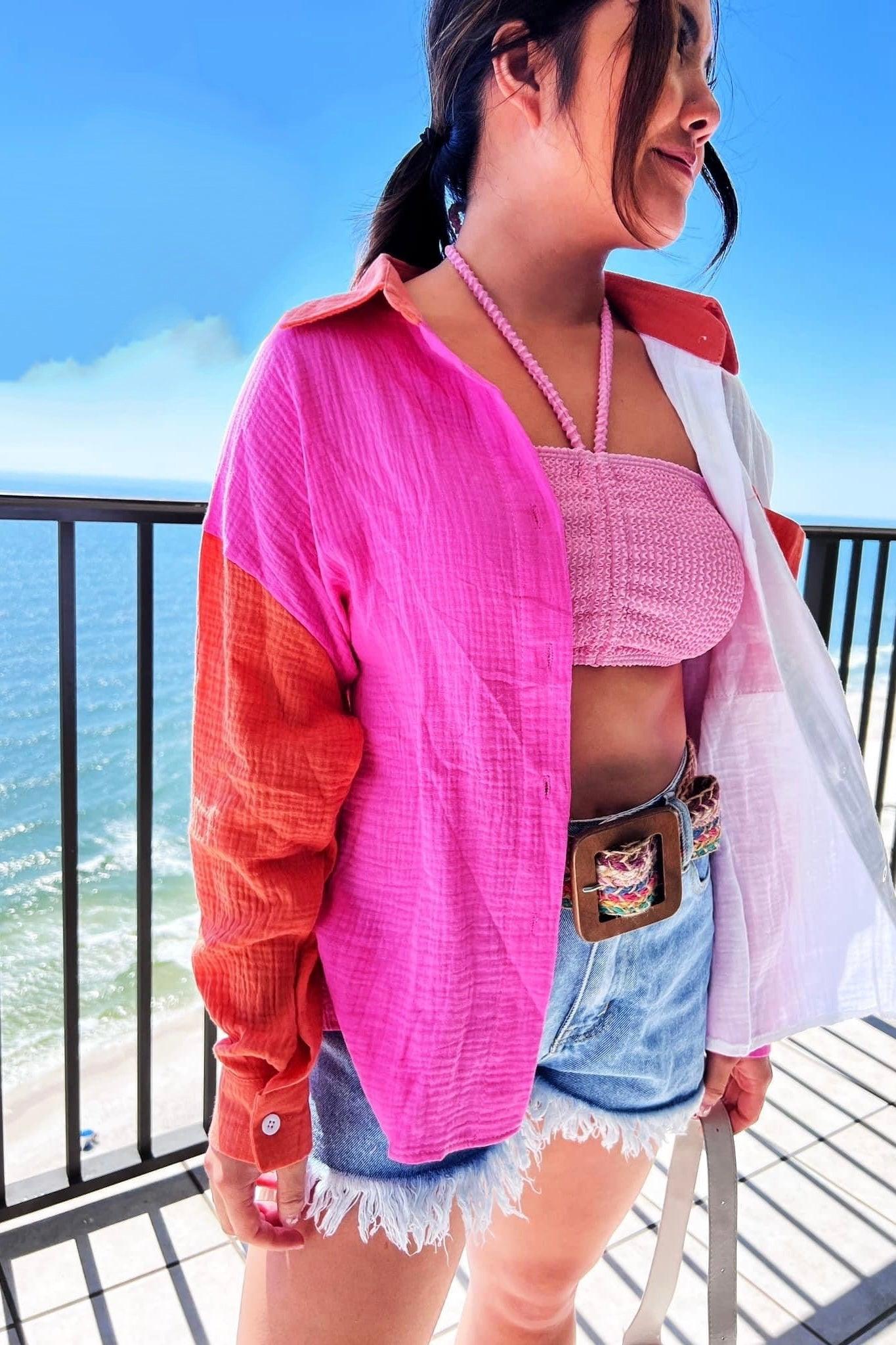 Woman wearing pink-orange-ivory color block shirt over pink knit crop top with denim shorts near ocean railing