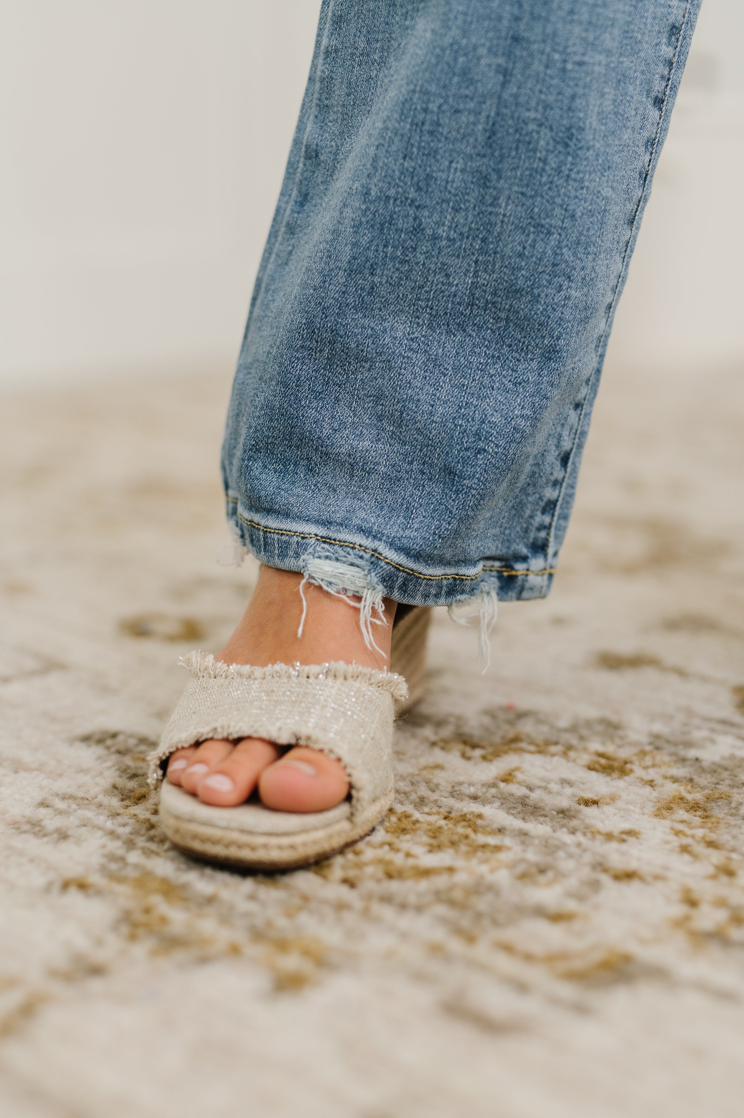 Close-up of frayed hem blue denim jeans paired with beige open-toe woven slip-on sandals on patterned carpet