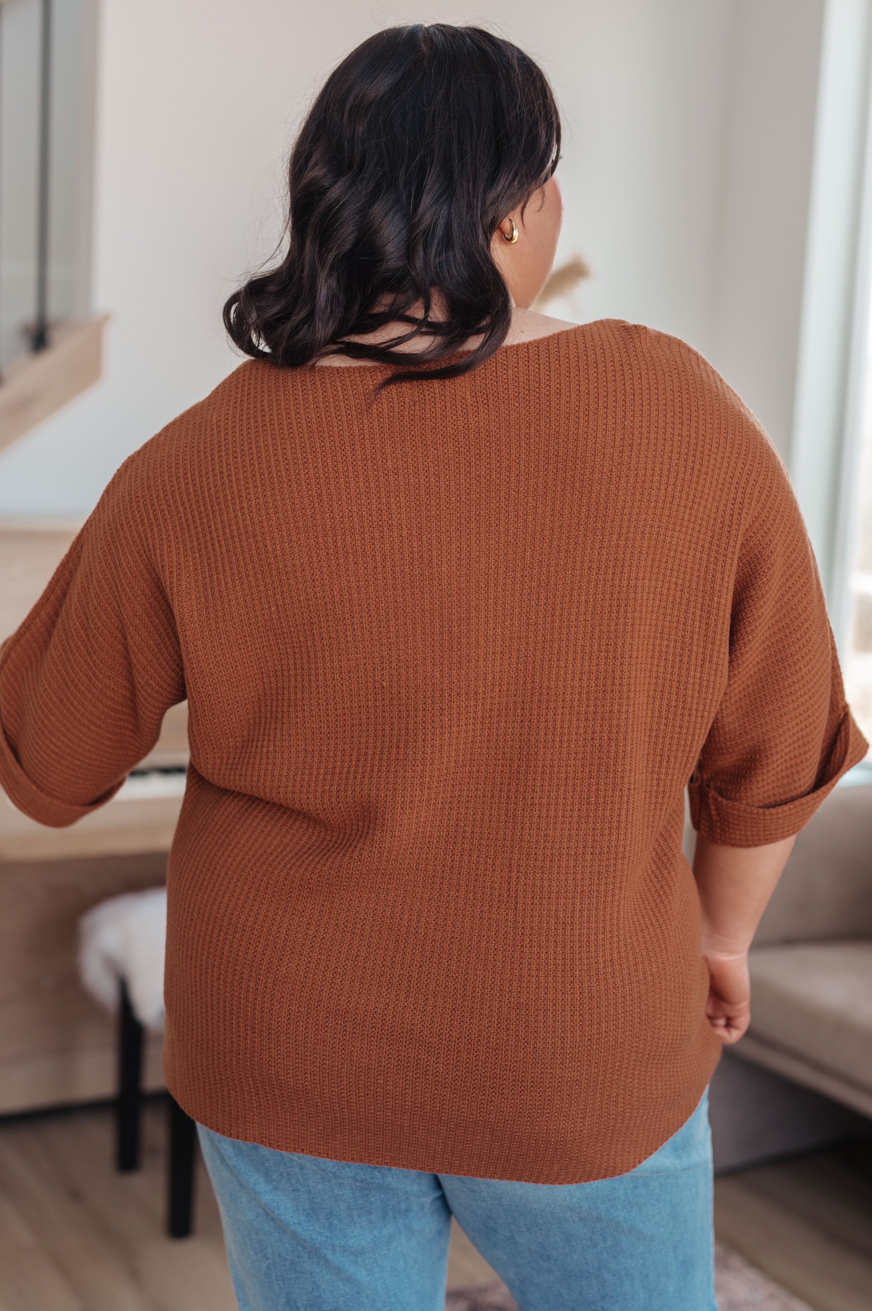 Woman seen from behind wearing a rust-colored textured knit top with rolled sleeves and light blue jeans indoors