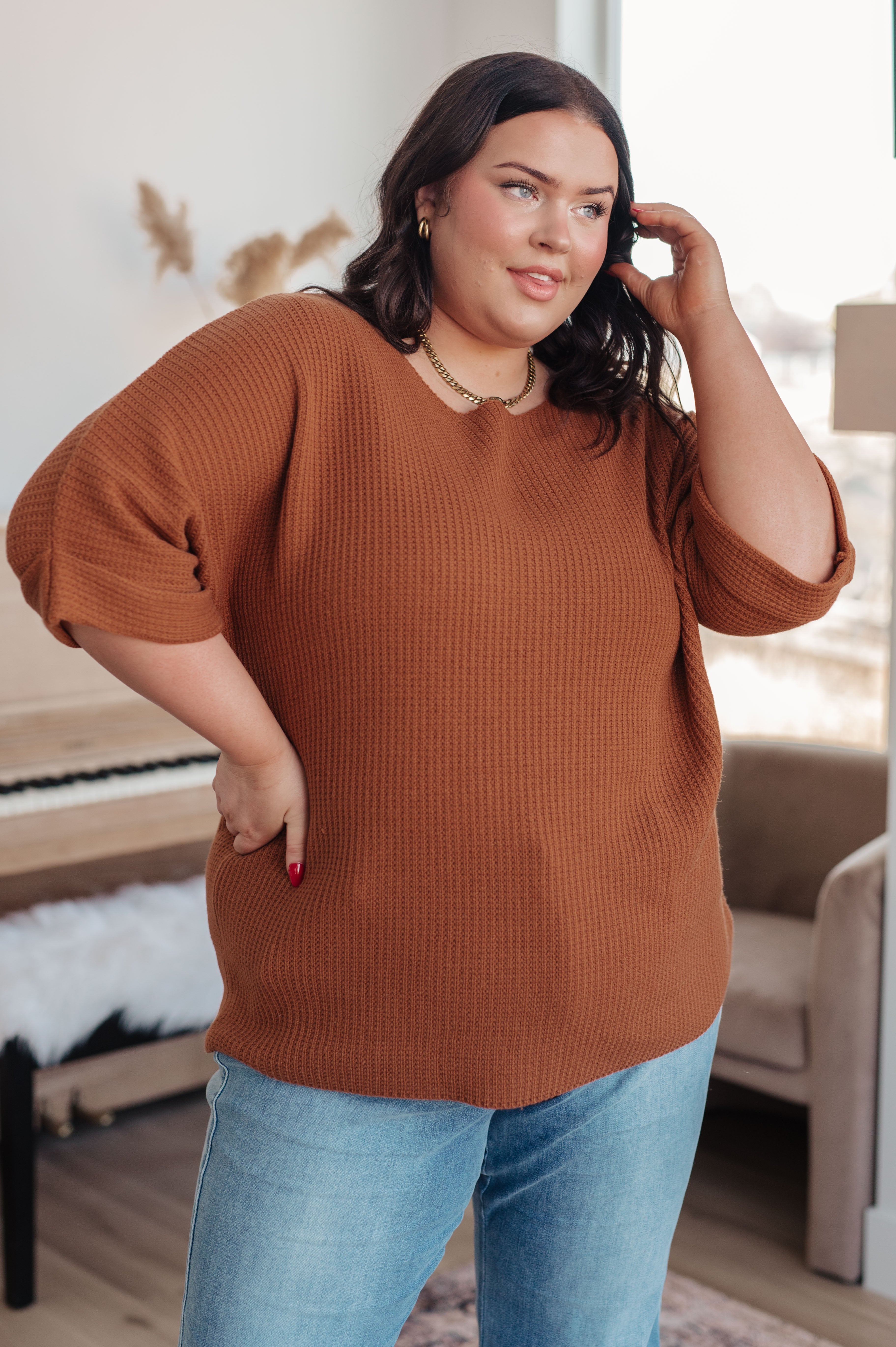 Plus-size woman wearing rust-colored knitted sweater top paired with light blue jeans standing indoors near window