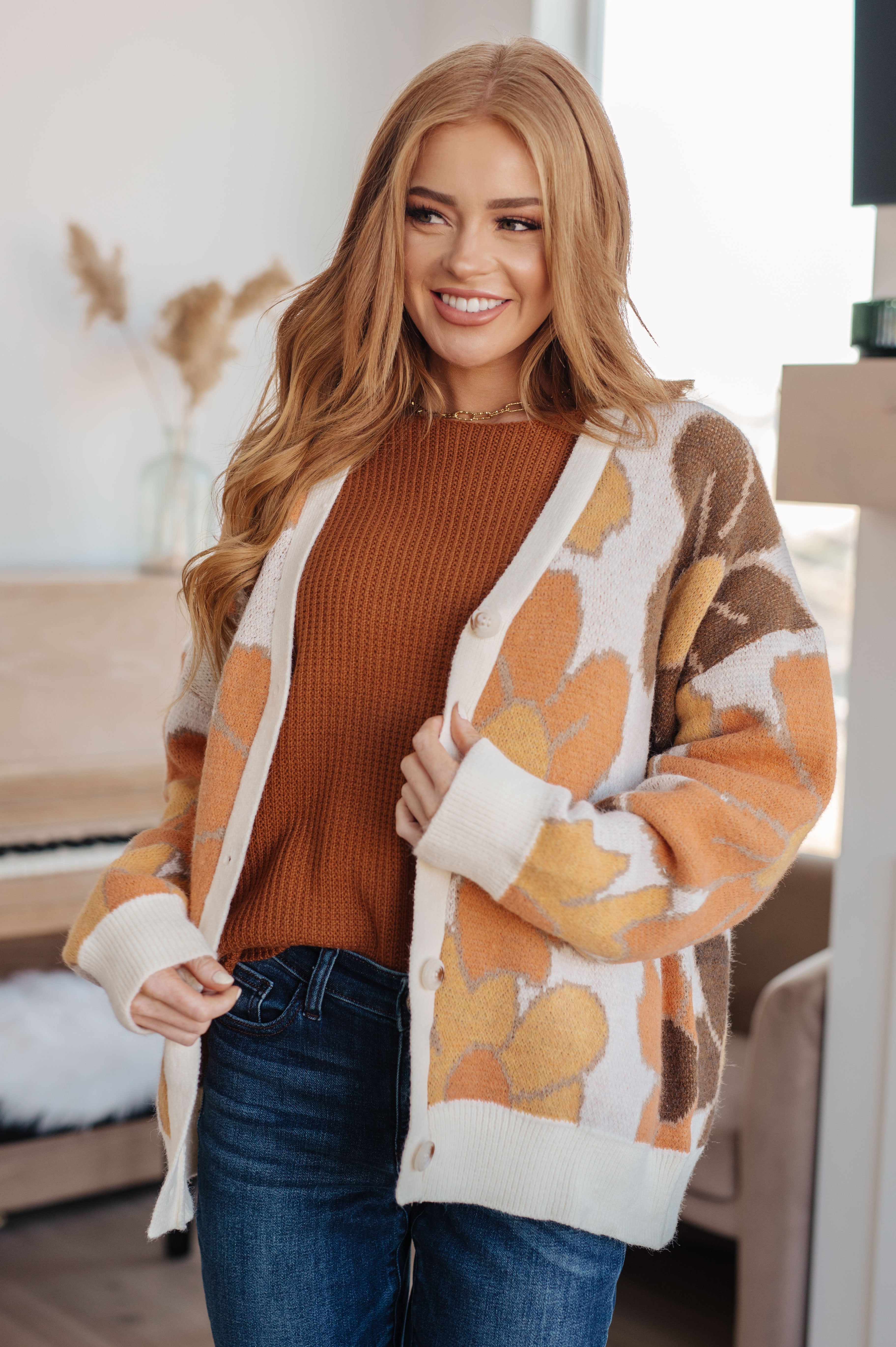 Smiling woman wearing rust knit top under oversized floral patterned cardigan paired with dark blue jeans indoors