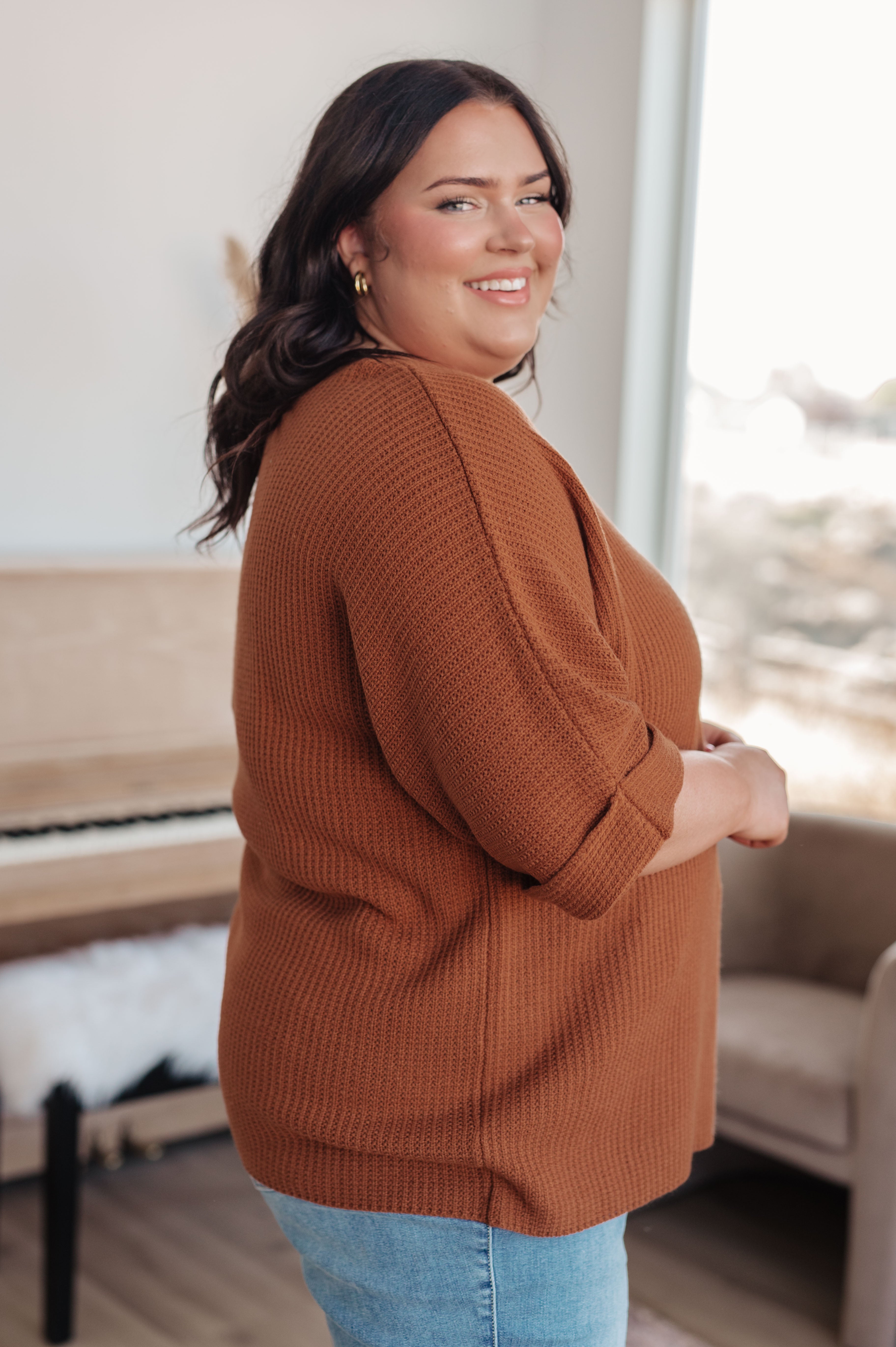 Curvy woman smiling indoors wearing a rust-colored textured knit top with rolled sleeves and light blue jeans
