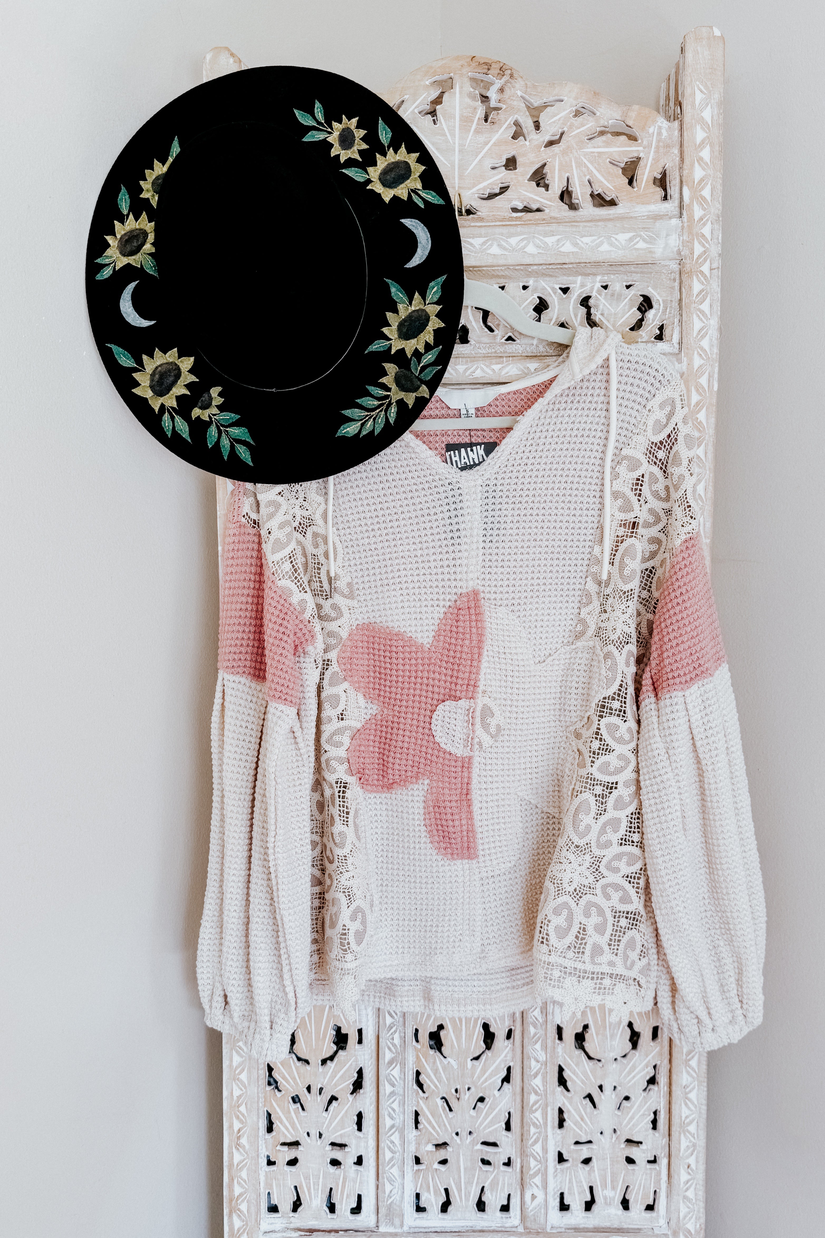 Cream waffle knit sweater with large pink flower and lace overlay hanging on carved white wooden panel with black sun and moon hat