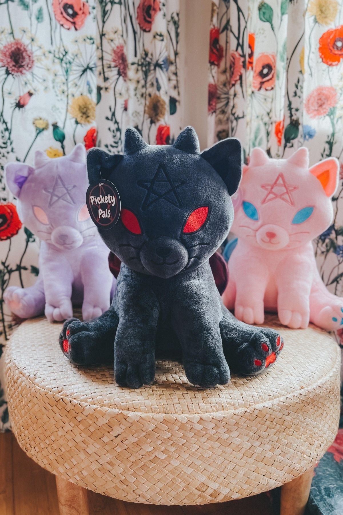 Charcoal black witchy kitten plush toy with red eyes pentagram detail and red paw accents seated on a woven stool with pastel plushies behind it