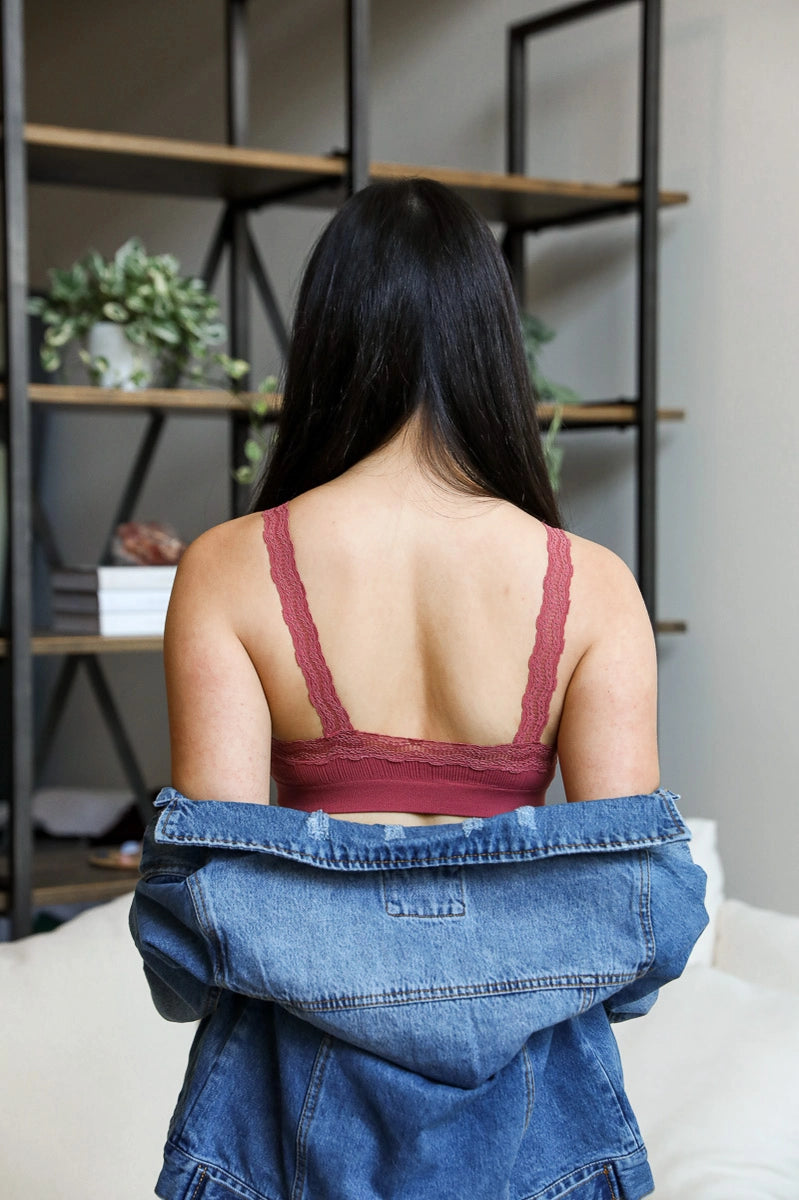 Woman with long dark hair wearing rust lace-trimmed bralette and slipping off blue denim jacket indoors by shelving unit