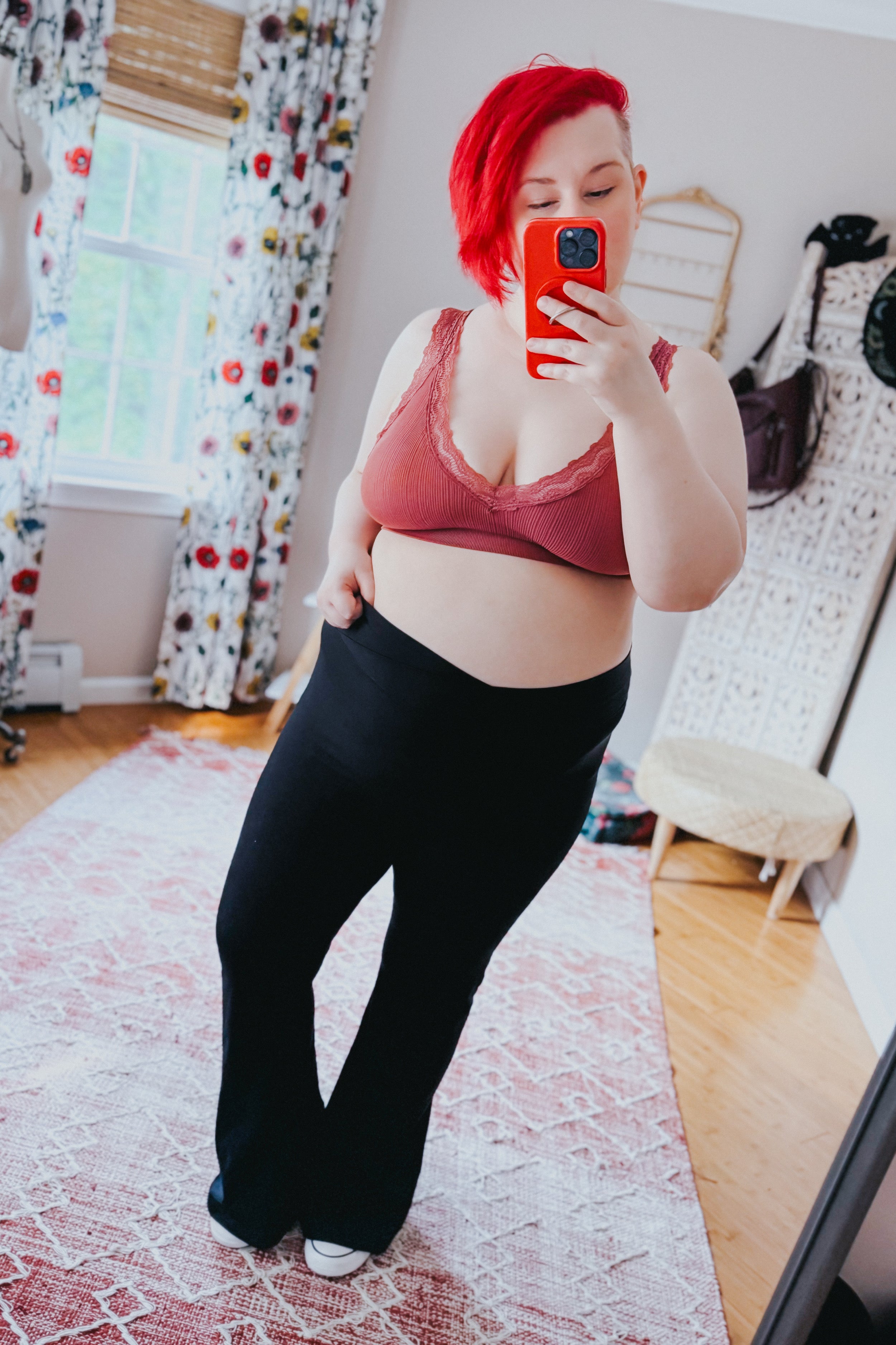 Curvy woman with bright red hair wearing rust-colored lace-trimmed bralette and black pants taking selfie in bright room